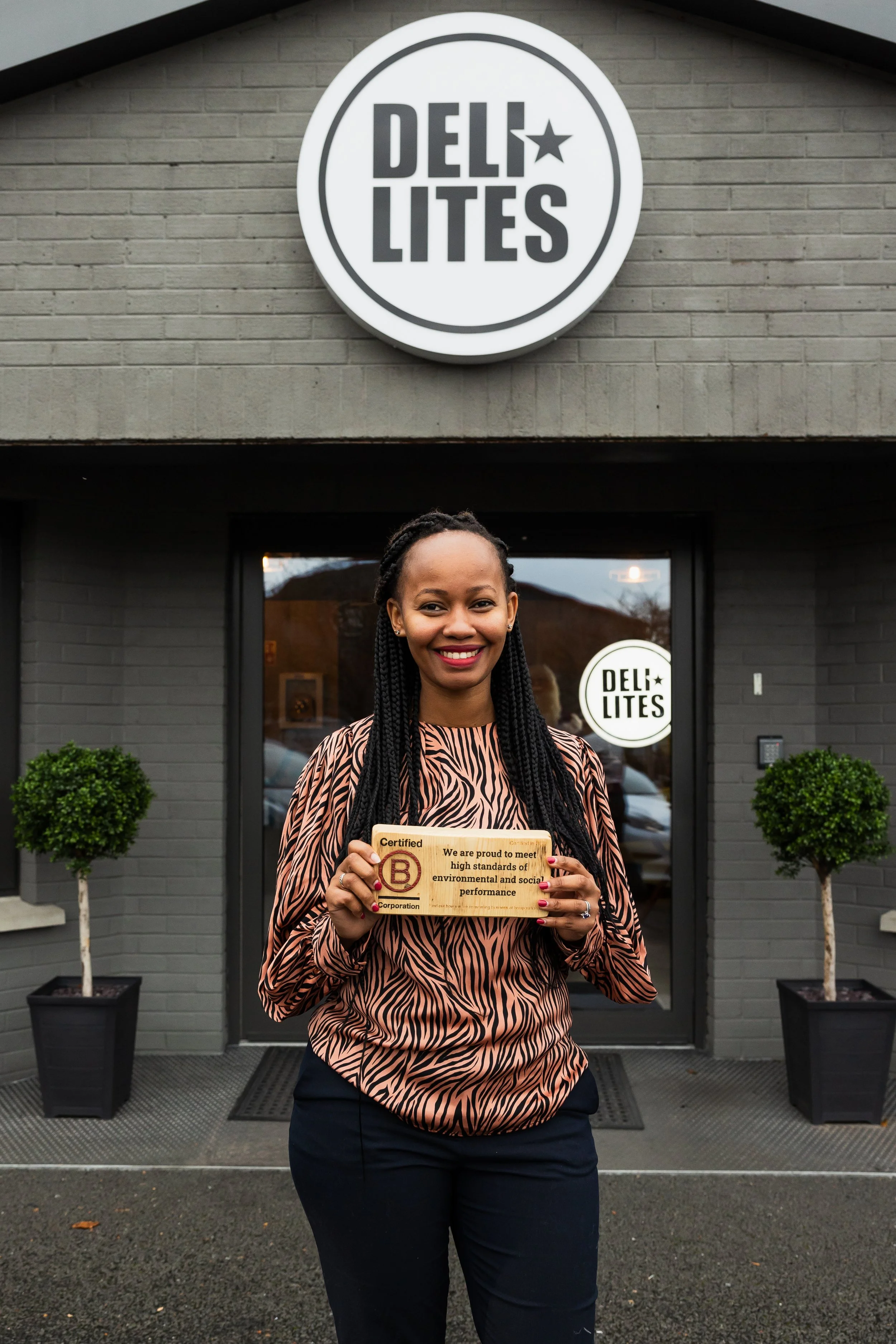 Woman standing in front of DELI LITES Factory, holding B-Corp certification plaque, smiling, with two potted plants on either side of the entrance.