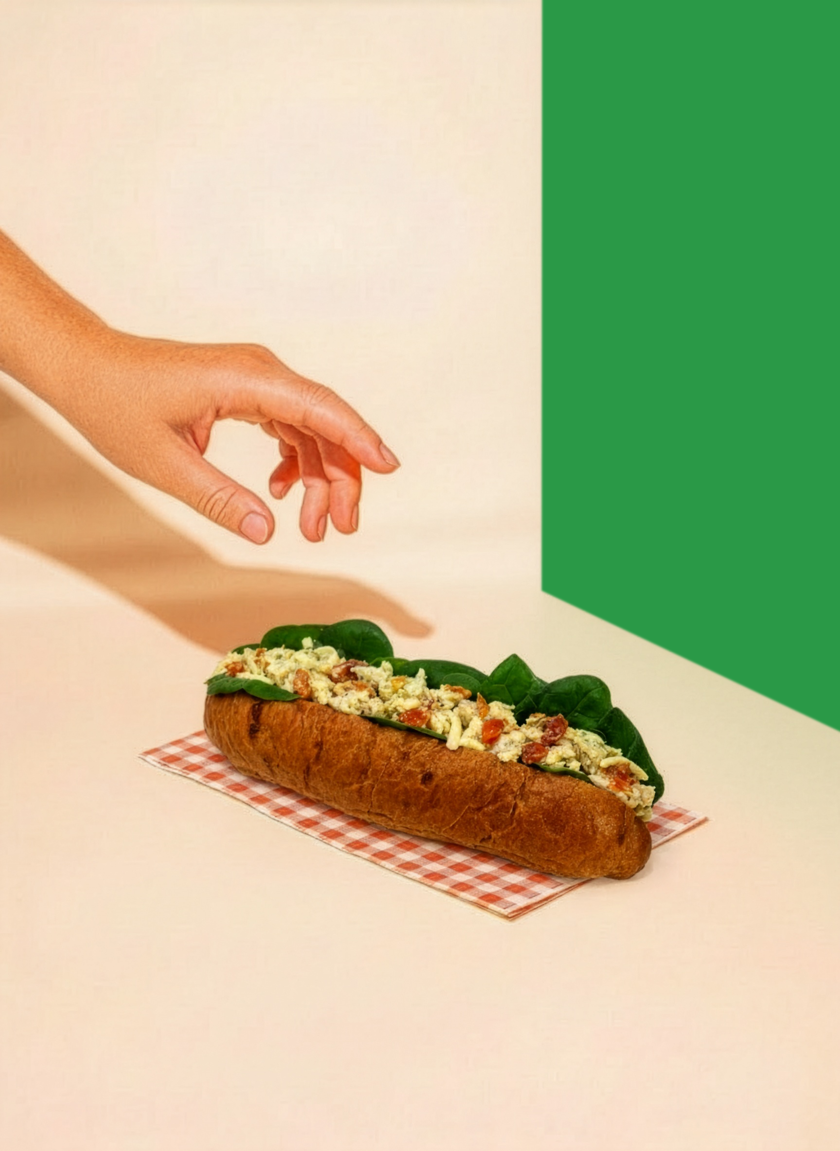 Hand reaching toward a sub sandwich on a checkered napkin with green spinach leaves and cheese and bacon inside.