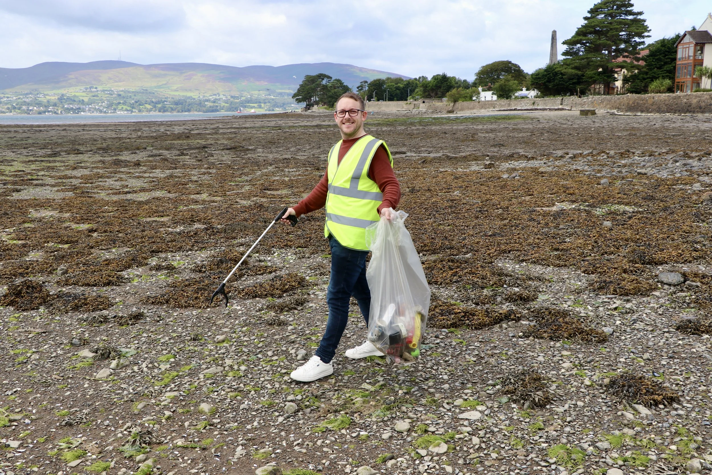 DELI LITES man in a yellow reflective vest picking up trash on a rocky shoreline with a garbage bag in hand, Mourne mountains in the background.