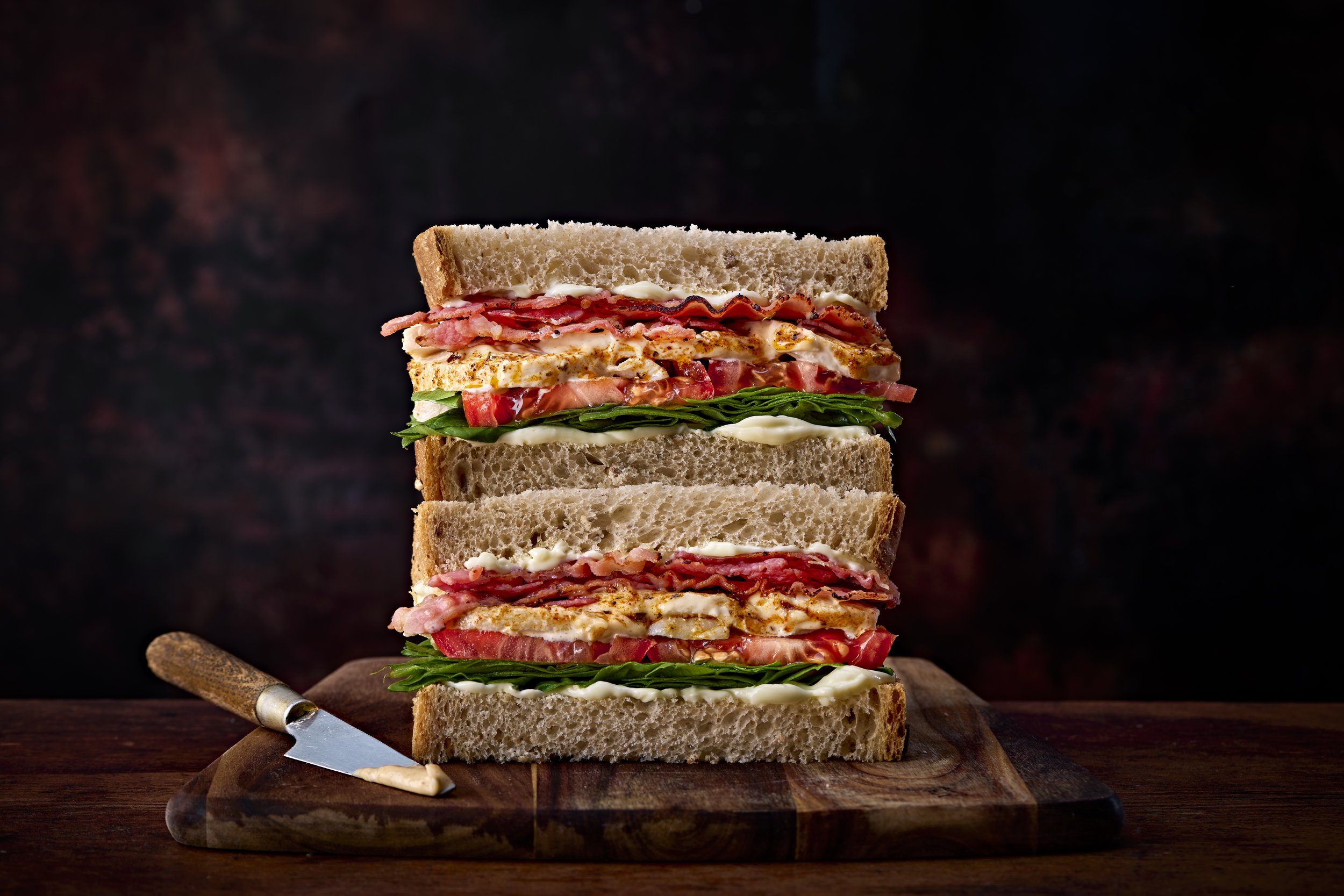 Stacked club sandwich with turkey, bacon, lettuce, tomato, and mayonnaise on whole wheat bread, placed on a wooden cutting board with a butter knife.
