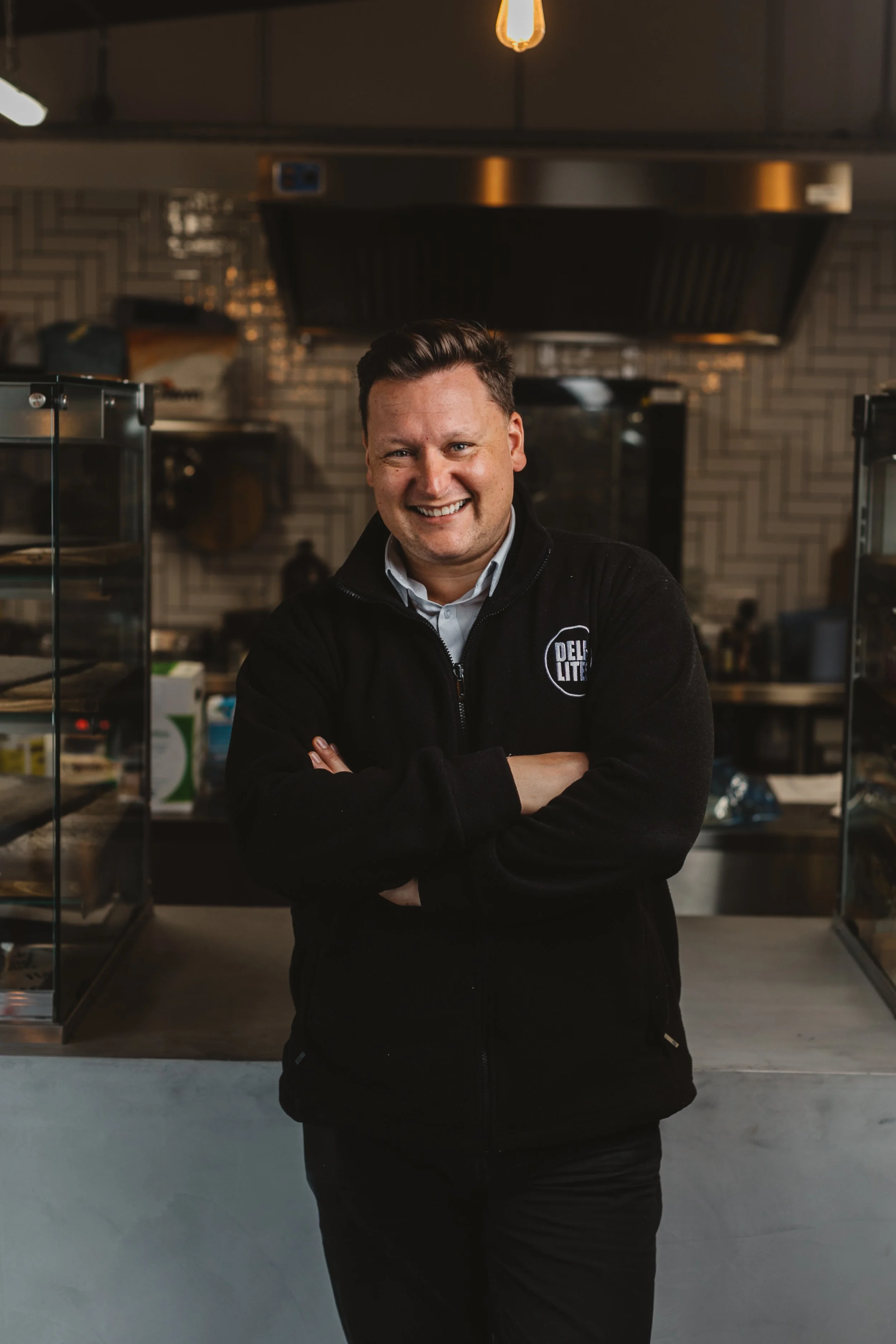 A smiling man with crossed arms standing inside a bakery or deli, wearing a black jacket with a logo, in front of a display case with baked goods, with a modern kitchen background.