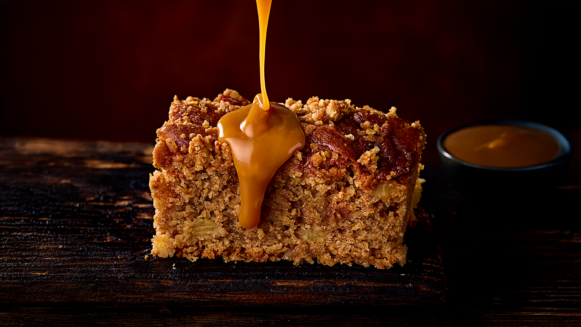 A piece of apple crumble cake with caramel sauce being poured over it, served on a dark wooden surface with a small bowl of caramel sauce in the background.