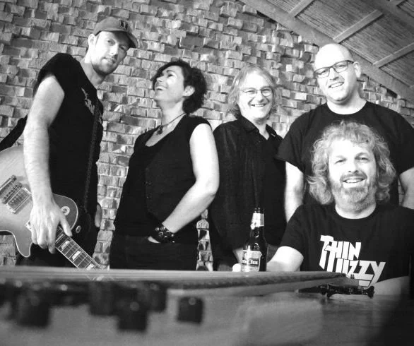 Five people in a casual setting, two men and three women, posing together with beverages on a table in front of a brick wall, some smiling and enjoying each other's company.