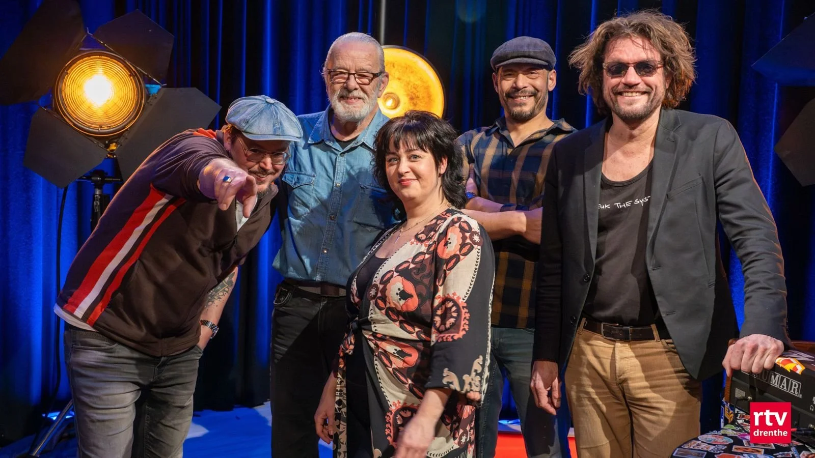 A group of five people posing for a photo on a stage with blue curtains, stage lights, and a yellow background. They are smiling and appear to be a comedy or performance group.