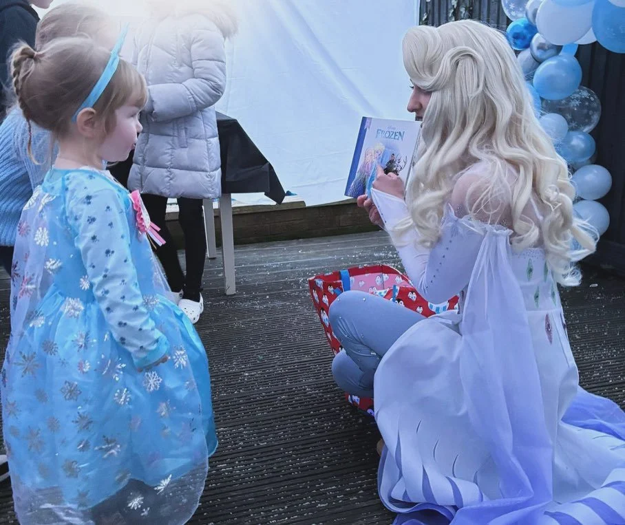 A woman dressed as Elsa from Frozen is kneeling and reading a book to a young girl dressed as a princess at a Frozen-themed birthday party with blue and white balloons.