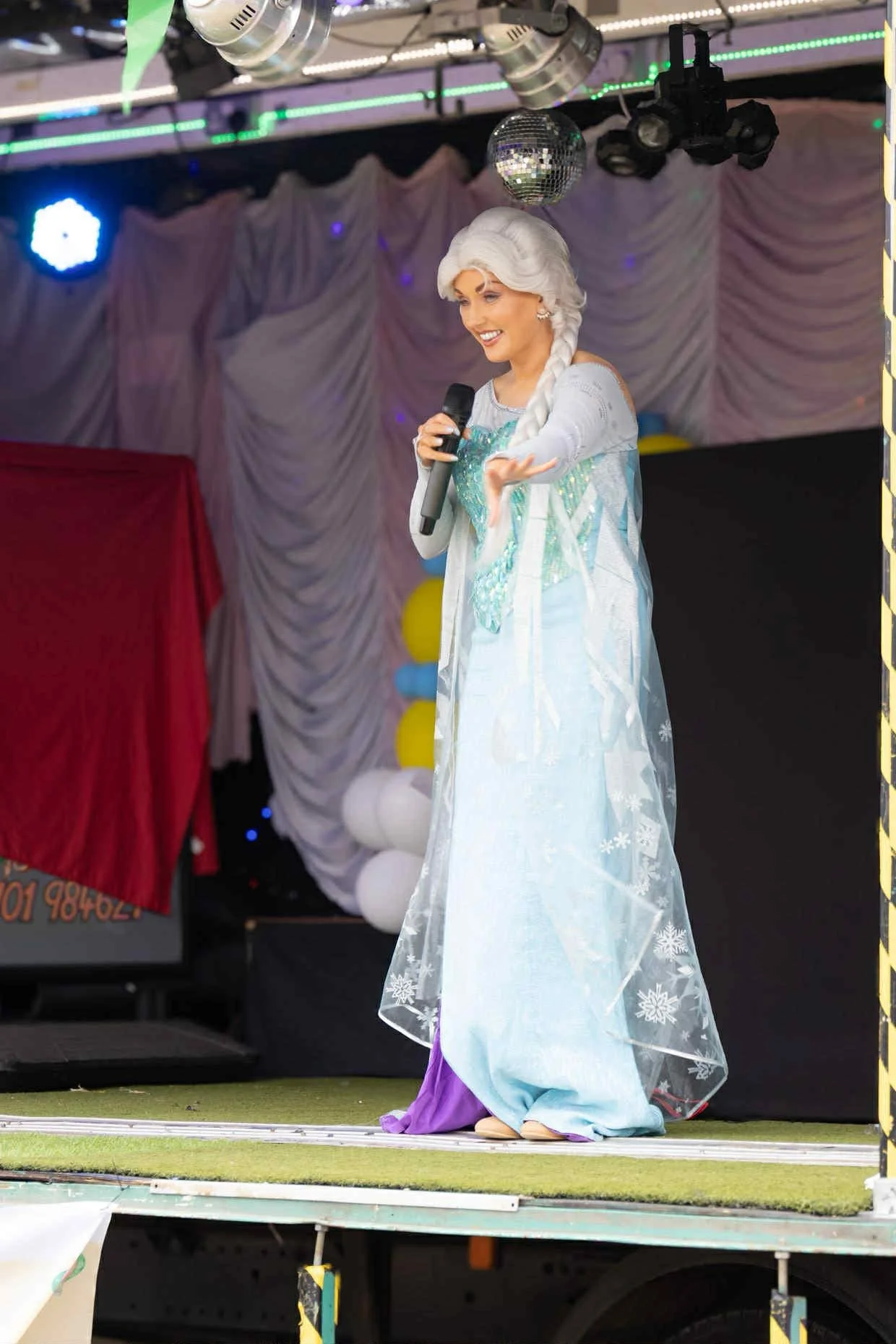 Woman dressed as Elsa from Frozen on stage, holding a microphone, with festive decorations and balloons in the background.