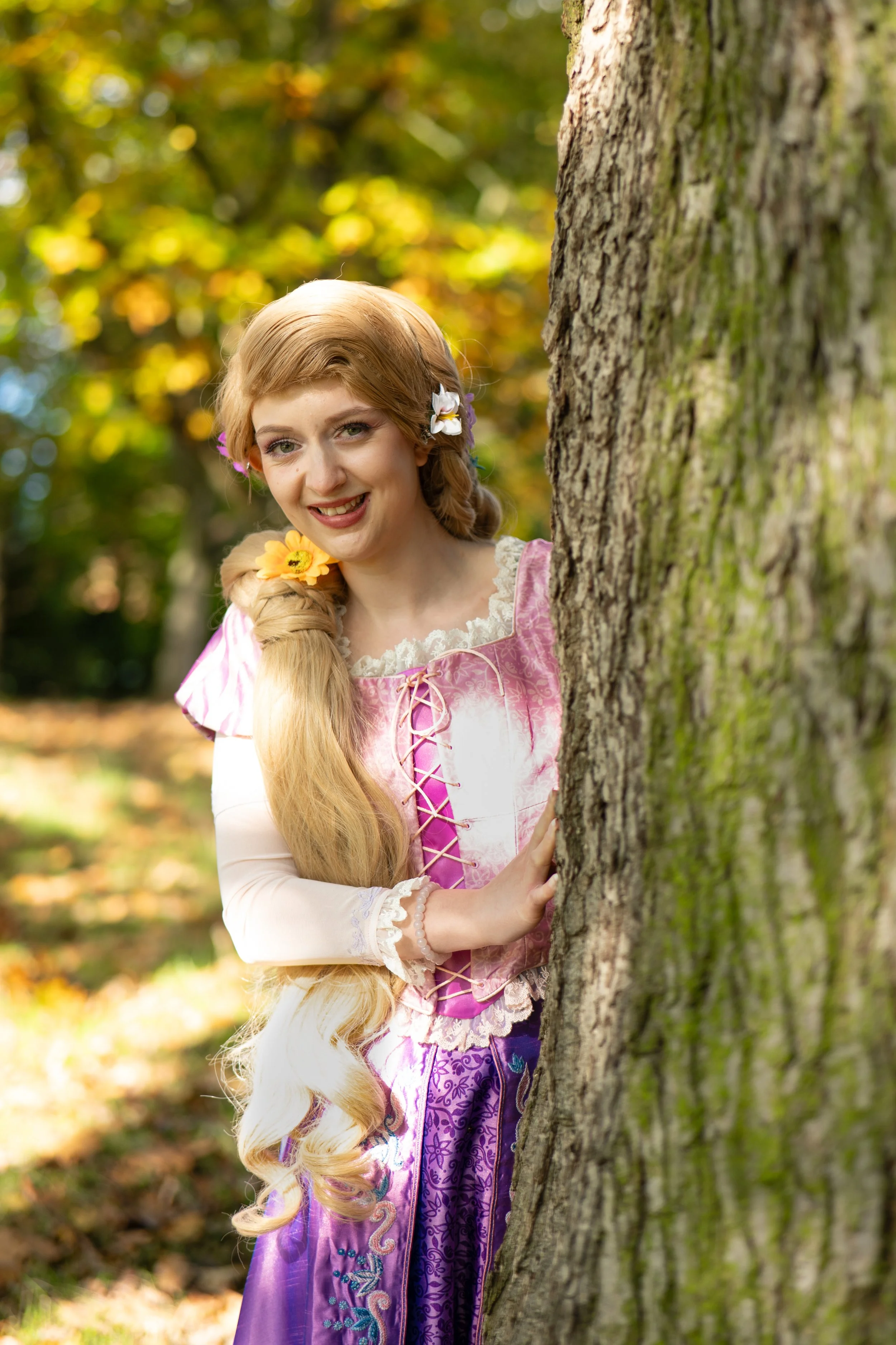 A woman dressed as Rapunzel in a pink and purple costume with long braided hair decorated with flowers, peeking out from behind a tree in a forest with autumn leaves.
