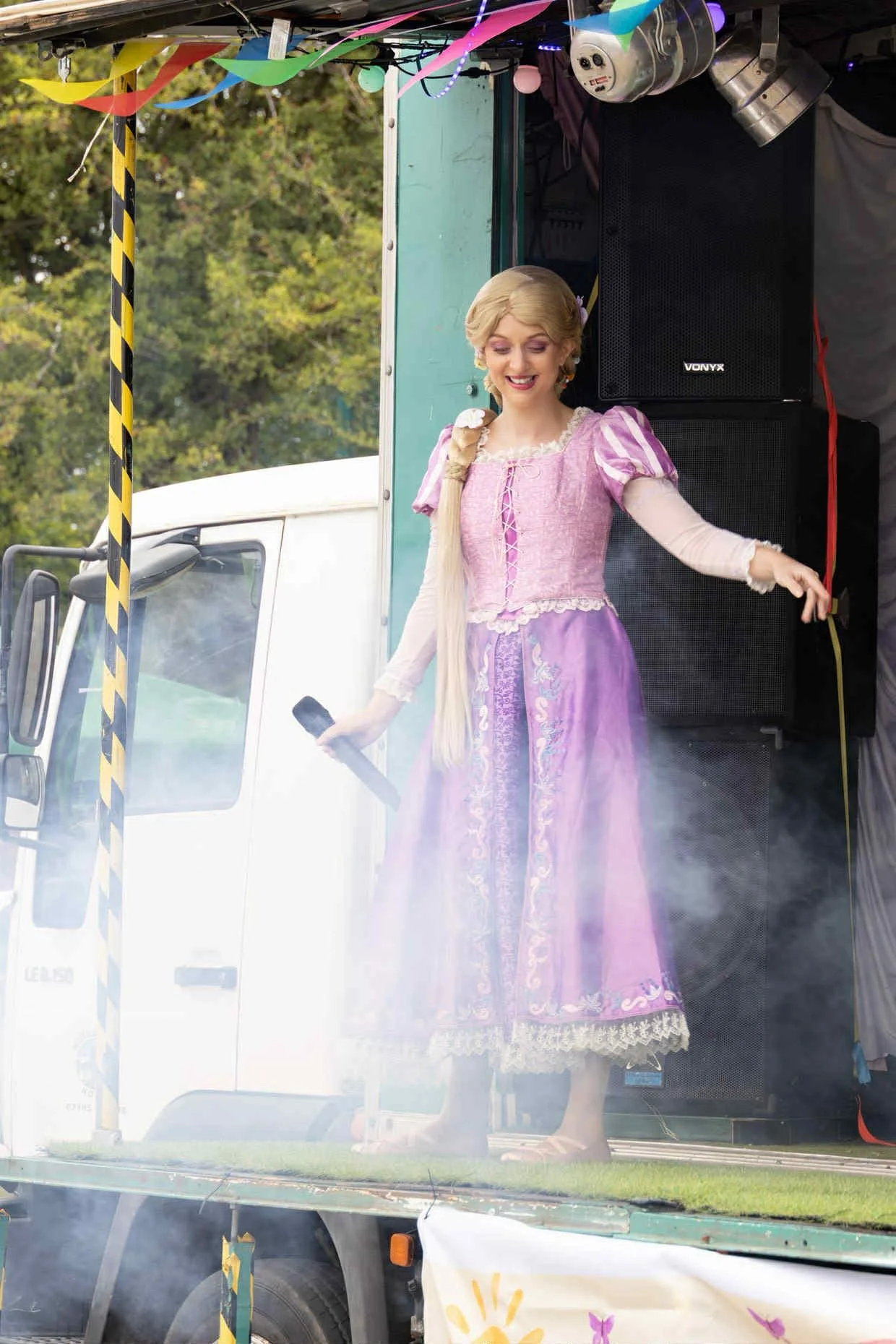 A woman dressed as Rapunzel from Disney's Tangled performing on a stage with a microphone in hand, standing in front of large speakers at an outdoor event. She has long blonde hair, wears a pink and purple dress, and is smiling.