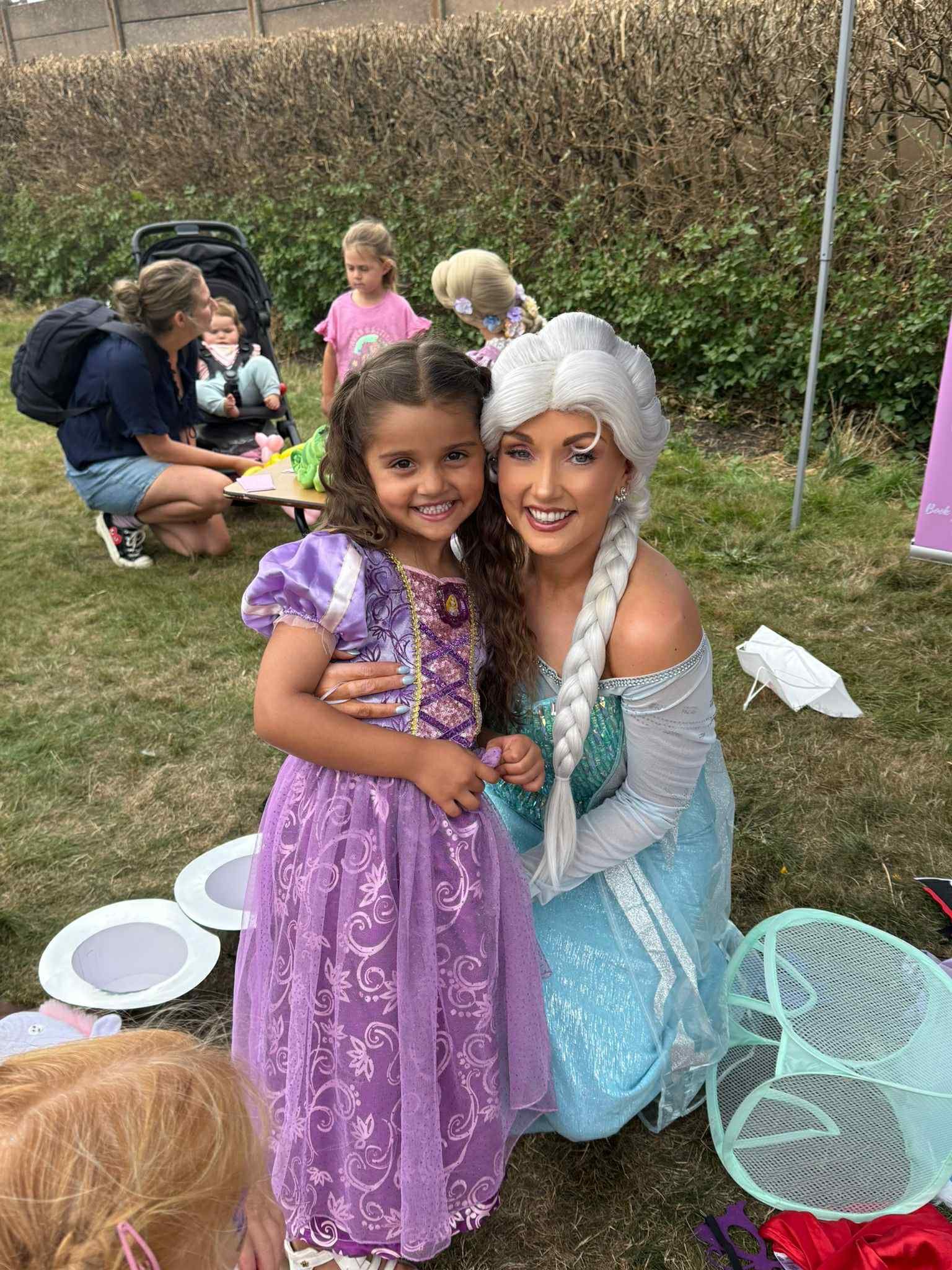 A young girl in a purple princess dress smiling and hugging a woman dressed as Elsa from Frozen, with several children and a woman with a backpack in the background.