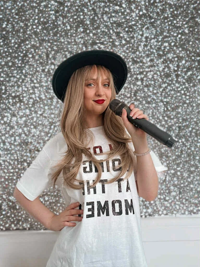 A woman with long blonde hair, wearing a black wide-brimmed hat and red lipstick, holding a microphone and smiling. She is dressed in a white t-shirt with bold black and red text. The background features a sparkling, glittery silver wall.