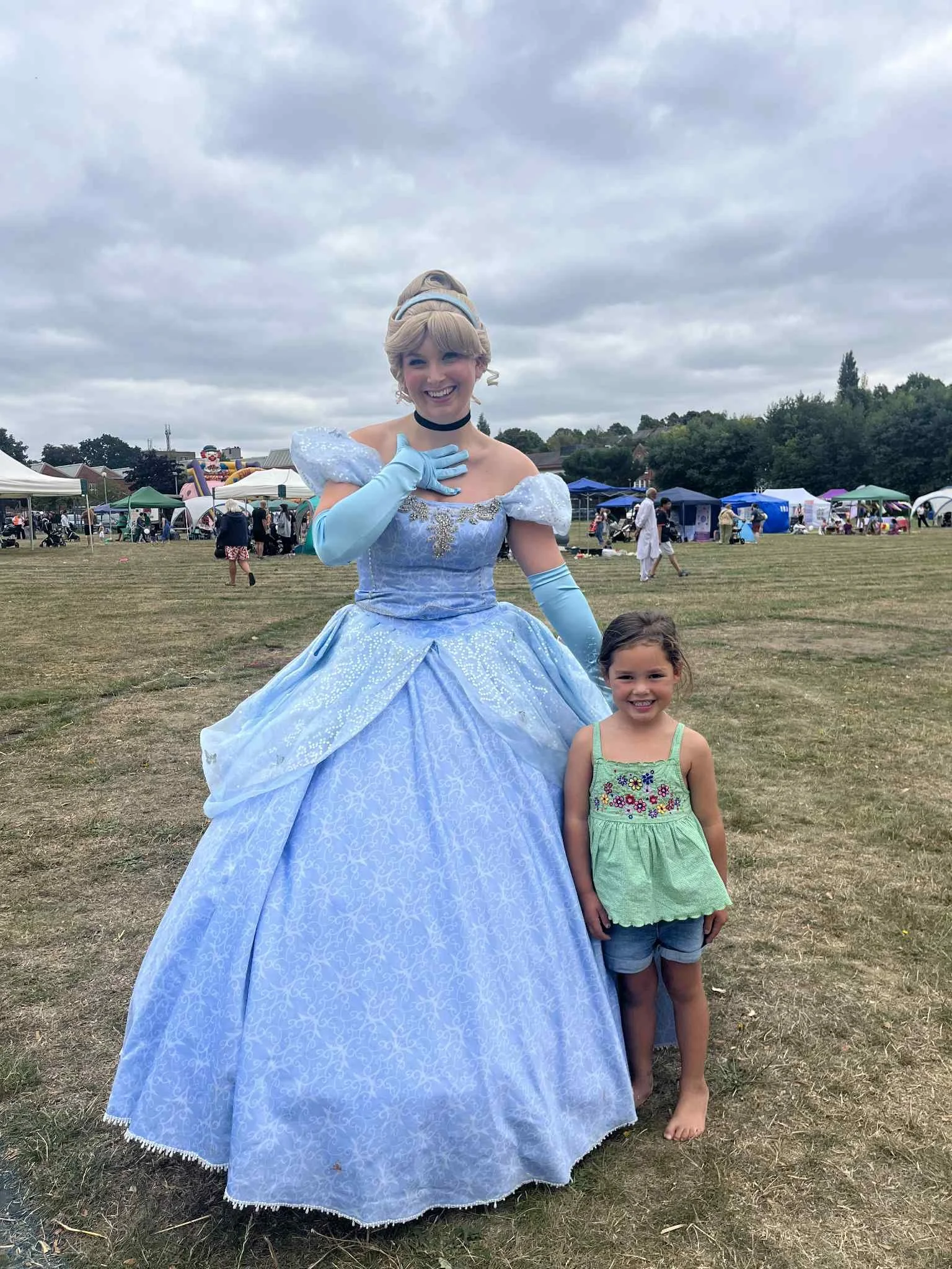 A woman dressed as a princess in a blue gown and a young girl in a green top and shorts standing on a grassy field during a fair or festival.