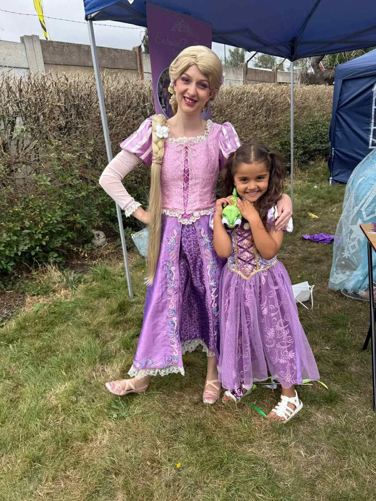 A woman dressed as Rapunzel from Disney's Tangled stands outdoors with a young girl holding a plush toy. Both are smiling and wearing purple costumes, with tents and bushes in the background.