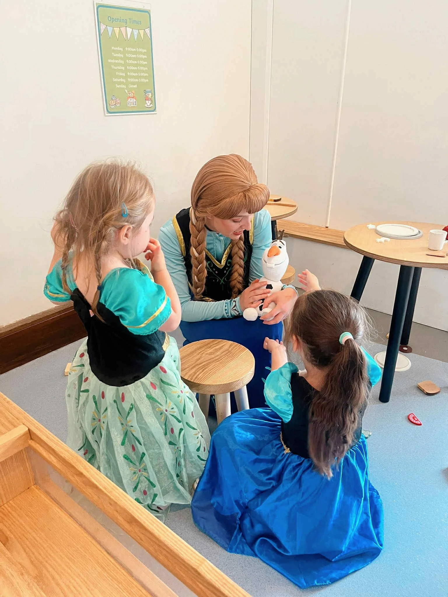 A woman dressed as Anna from Frozen holding Olaf puppet, with two young girls in princess costumes sitting and standing around her, engaged in a pretend play with Olaf, in a room with tables and a sign on the wall showing opening times.