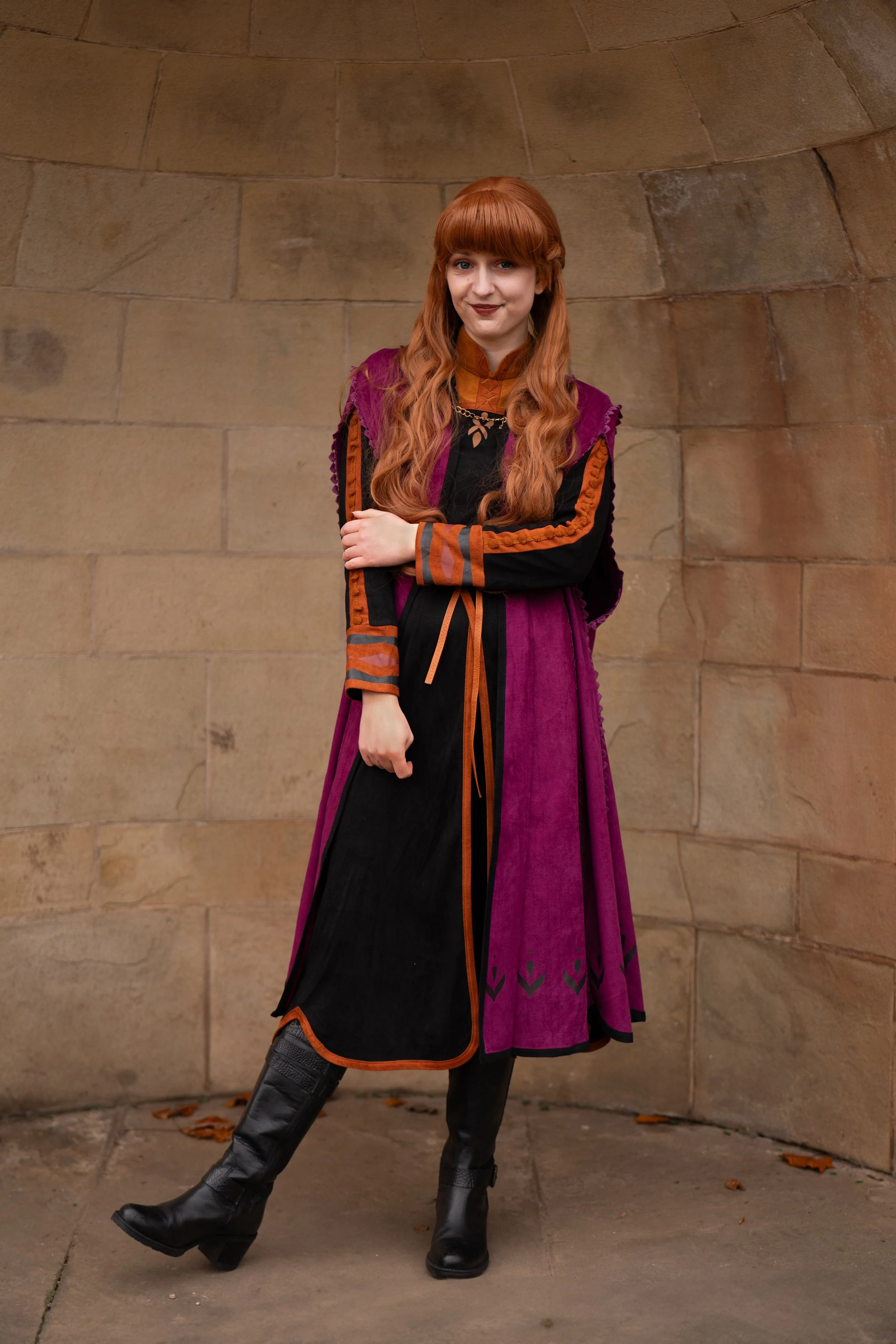A woman with long red hair wearing a costume inspired by Disney's Anna from Frozen, standing outdoors against a tan stone wall.