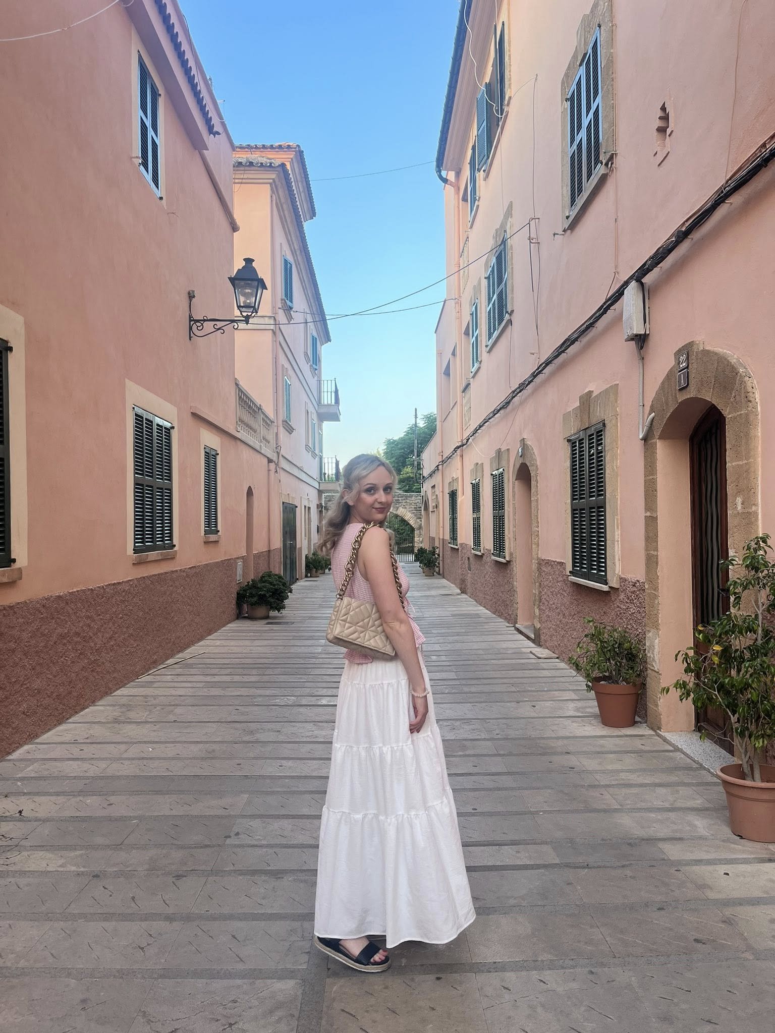 A woman with blonde hair wearing a pink top and long white skirt, carrying a beige quilted purse, standing on a narrow street with pastel pink buildings, black shutters, potted plants, and a clear blue sky.