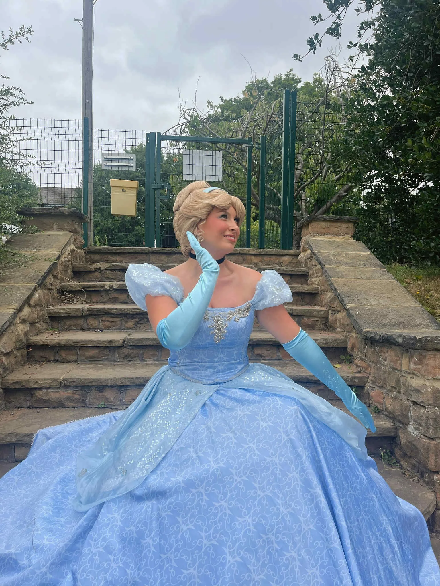 A woman dressed as Cinderella in a blue gown with puffy sleeves, long gloves, and a blonde updo hairstyle, sitting on stone steps outside, touching her face and smiling.
