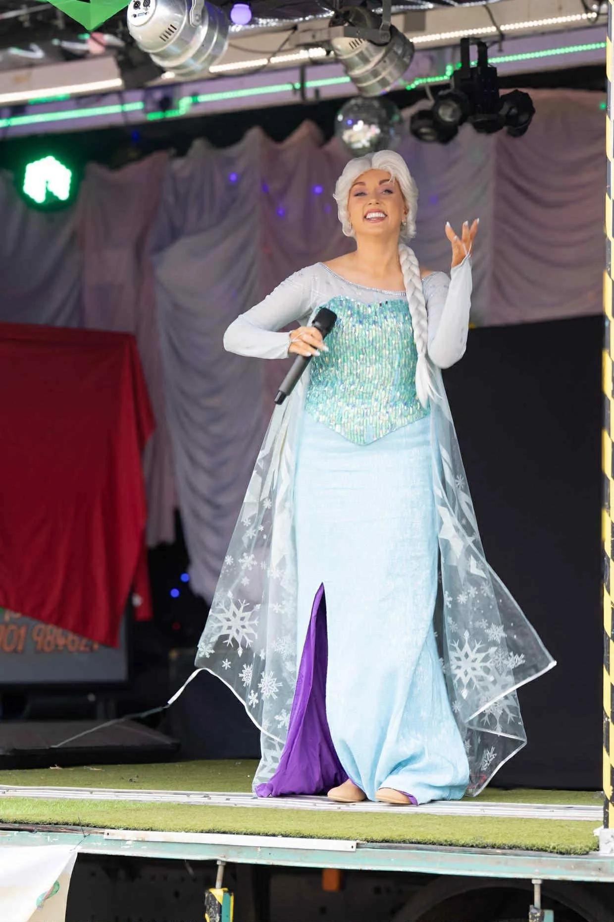 Person dressed as Elsa from Frozen performing on stage with a microphone in hand, smiling, with stage lights and curtains in the background.