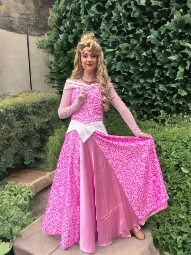 A woman dressed as Princess Aurora from Sleeping Beauty, standing outdoors on a stone path, surrounded by green bushes and trees.