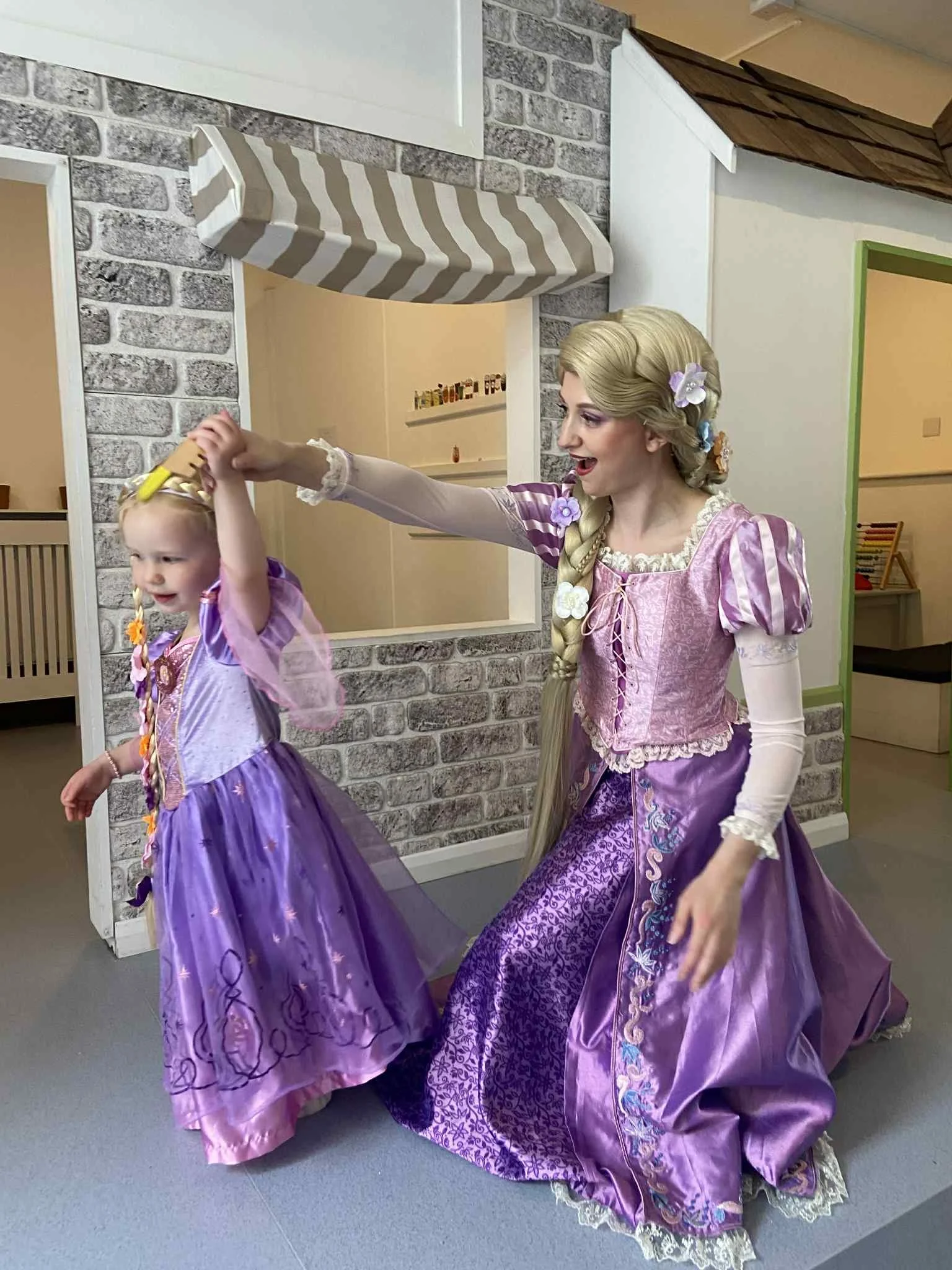 A woman dressed as Rapunzel from Disney's Tangled is dancing with a young girl in a purple Rapunzel dress inside a play area or indoor space.