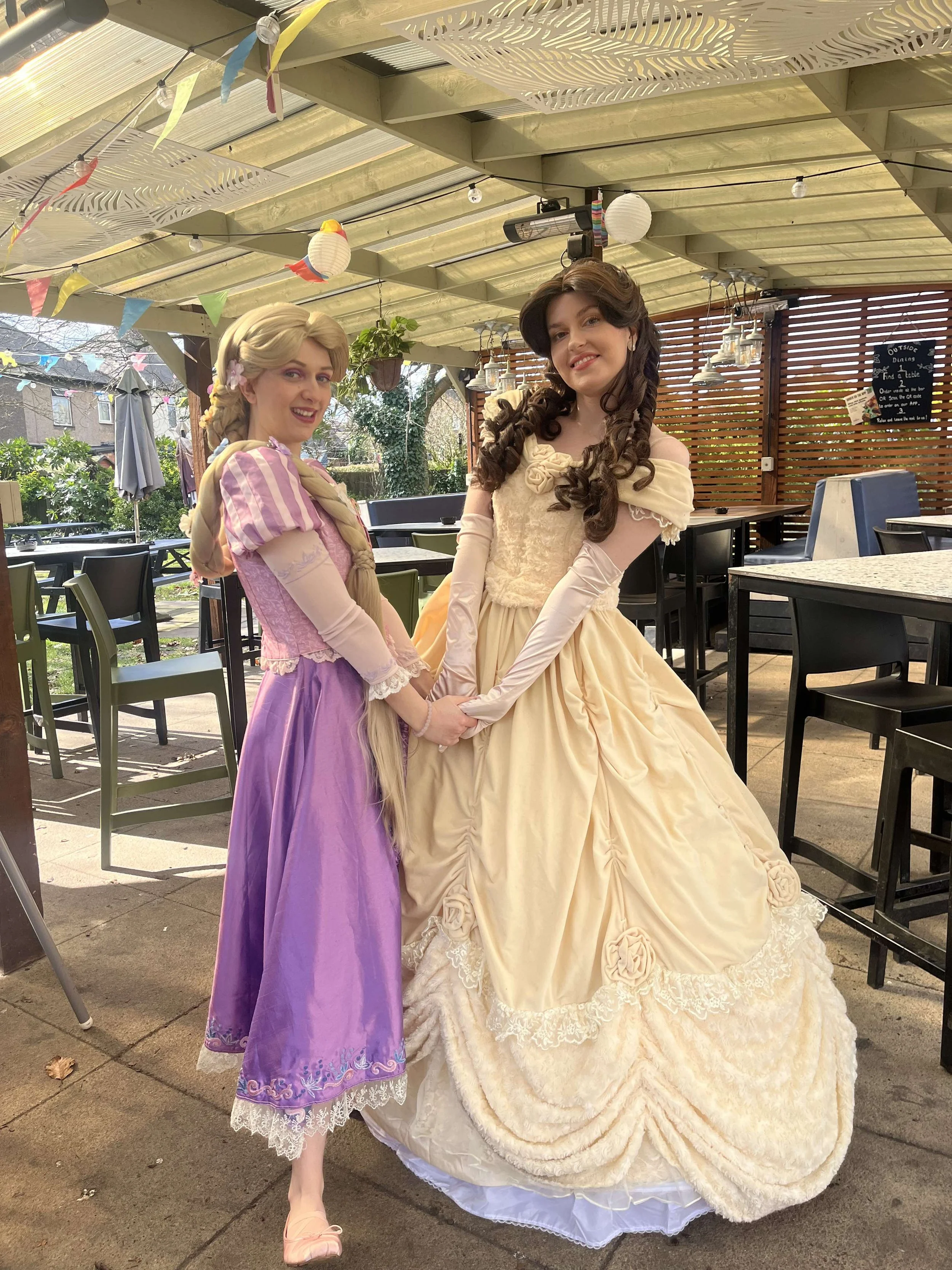 Two women dressed as Disney princesses Rapunzel and Belle holding hands and smiling, standing on a patio under a roof decorated with paper lanterns and bunting.