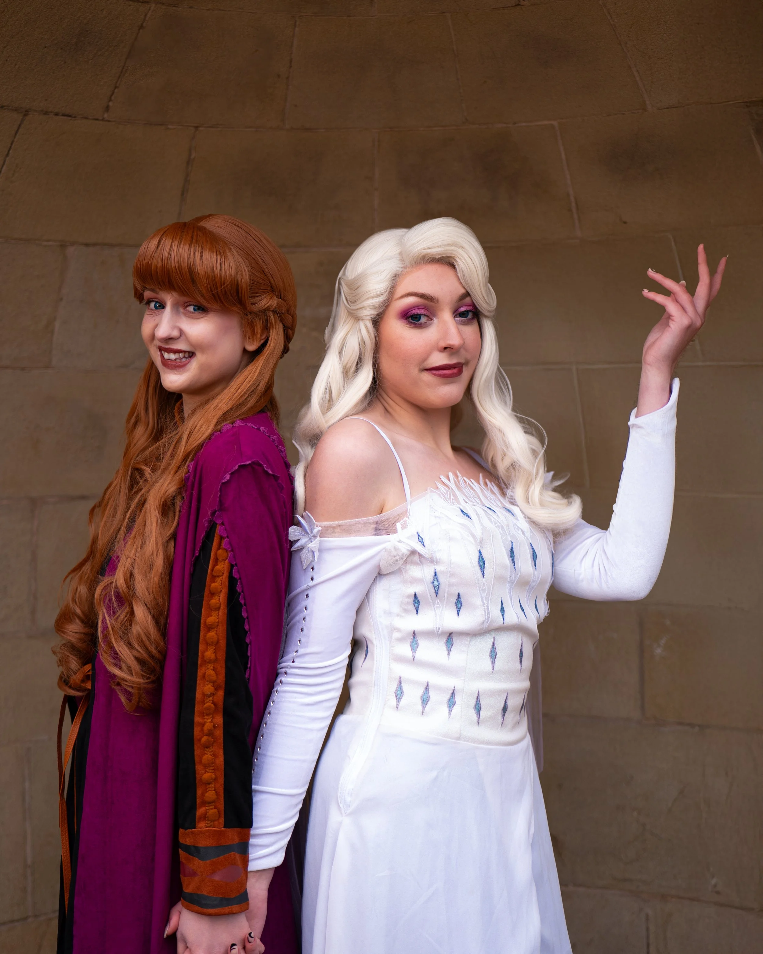 Two women, one with red hair in a purple dress and one with platinum blonde hair in a white dress, standing back-to-back against a stone wall, smiling and posing.
