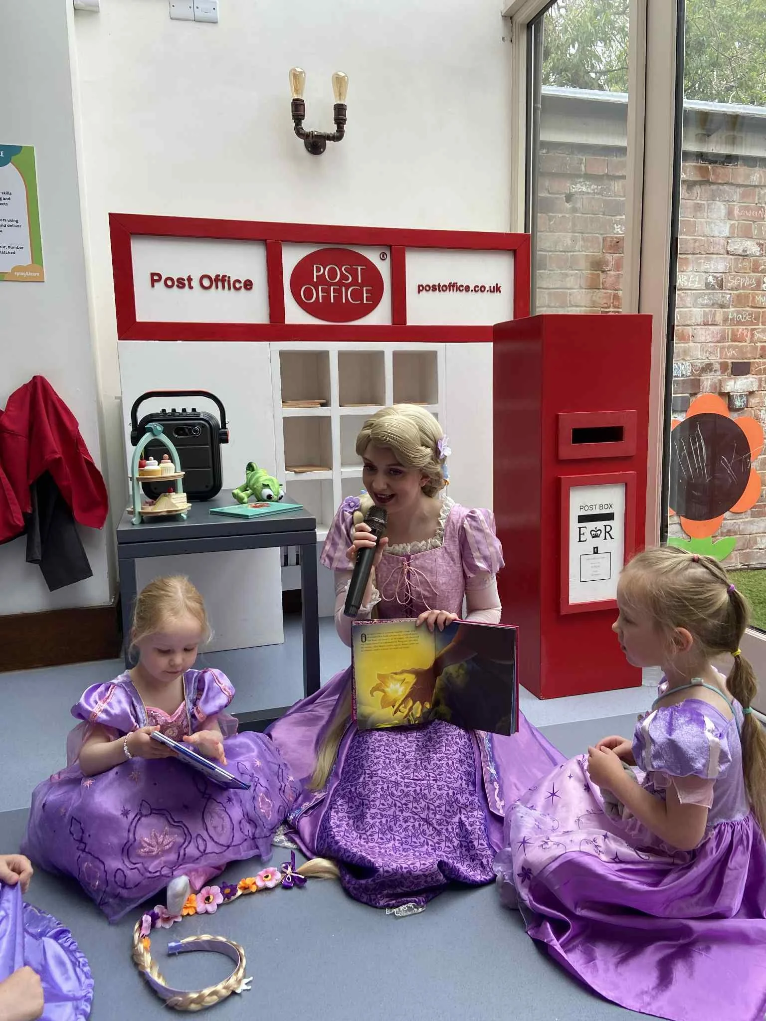 A woman dressed as Rapunzel sitting on the floor with three young girls also dressed as Rapunzel, reading a storybook together in a community space with a Disney-themed backdrop.