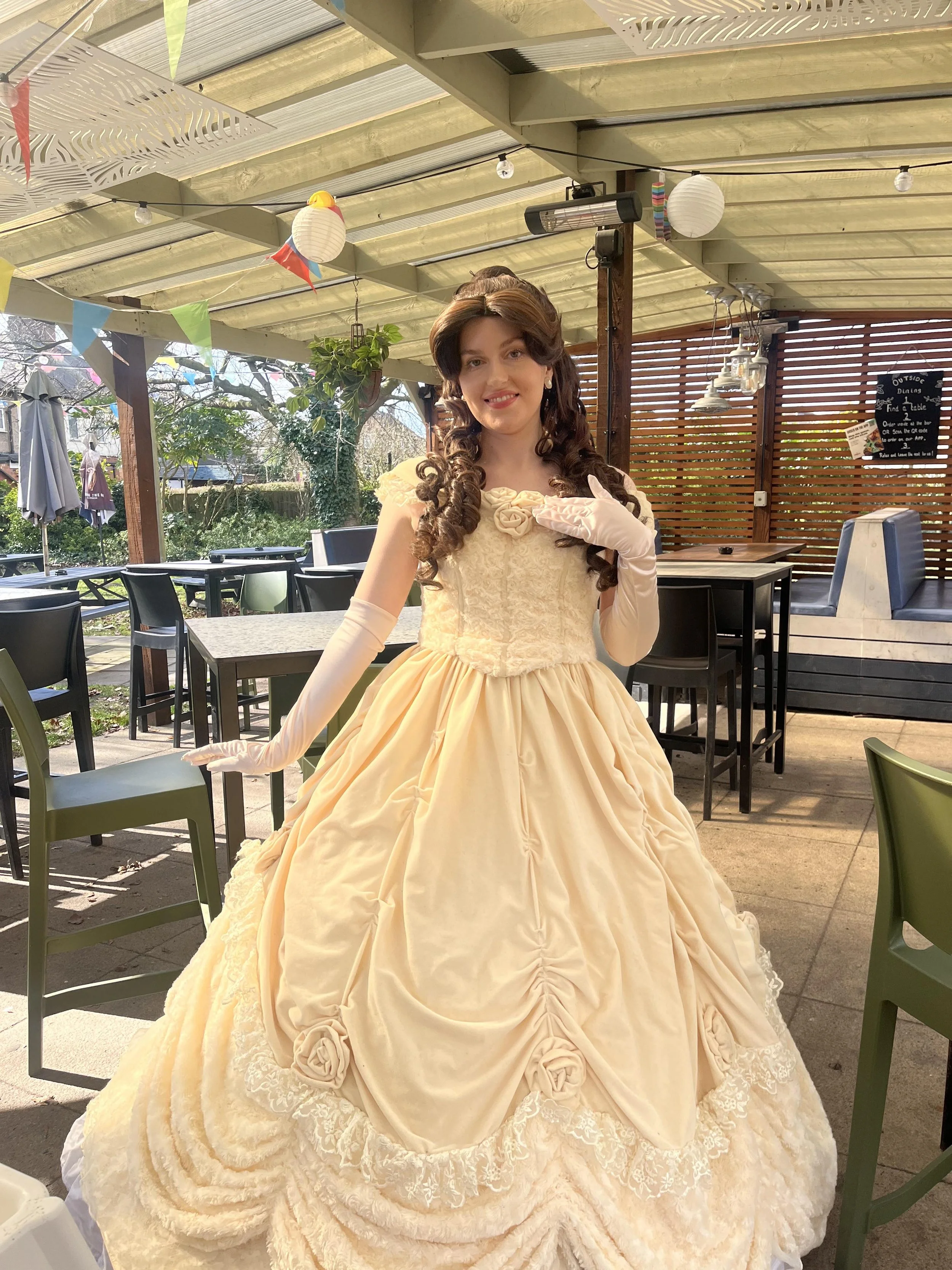 A woman dressed as Belle from Beauty and the Beast, wearing a vintage cream-colored gown with floral details, gloves, and a floral accessory, standing in a decorated patio area.