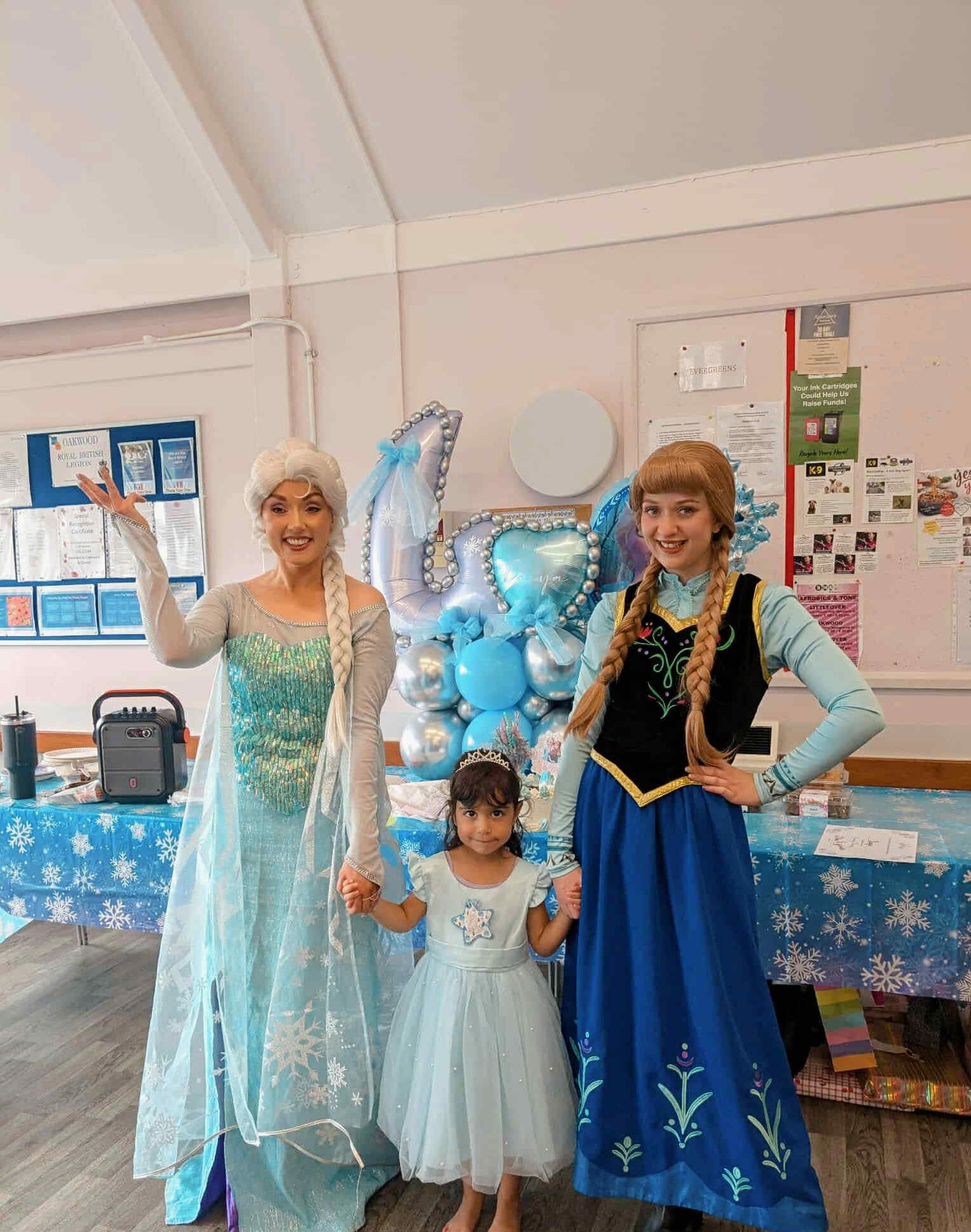 Two women dressed as Disney princesses, Elsa and Anna, holding hands with a young girl in a fairy-tale princess dress, standing in front of a balloon decoration with blue and silver balloons and a heart-shaped balloon, in a decorated room.