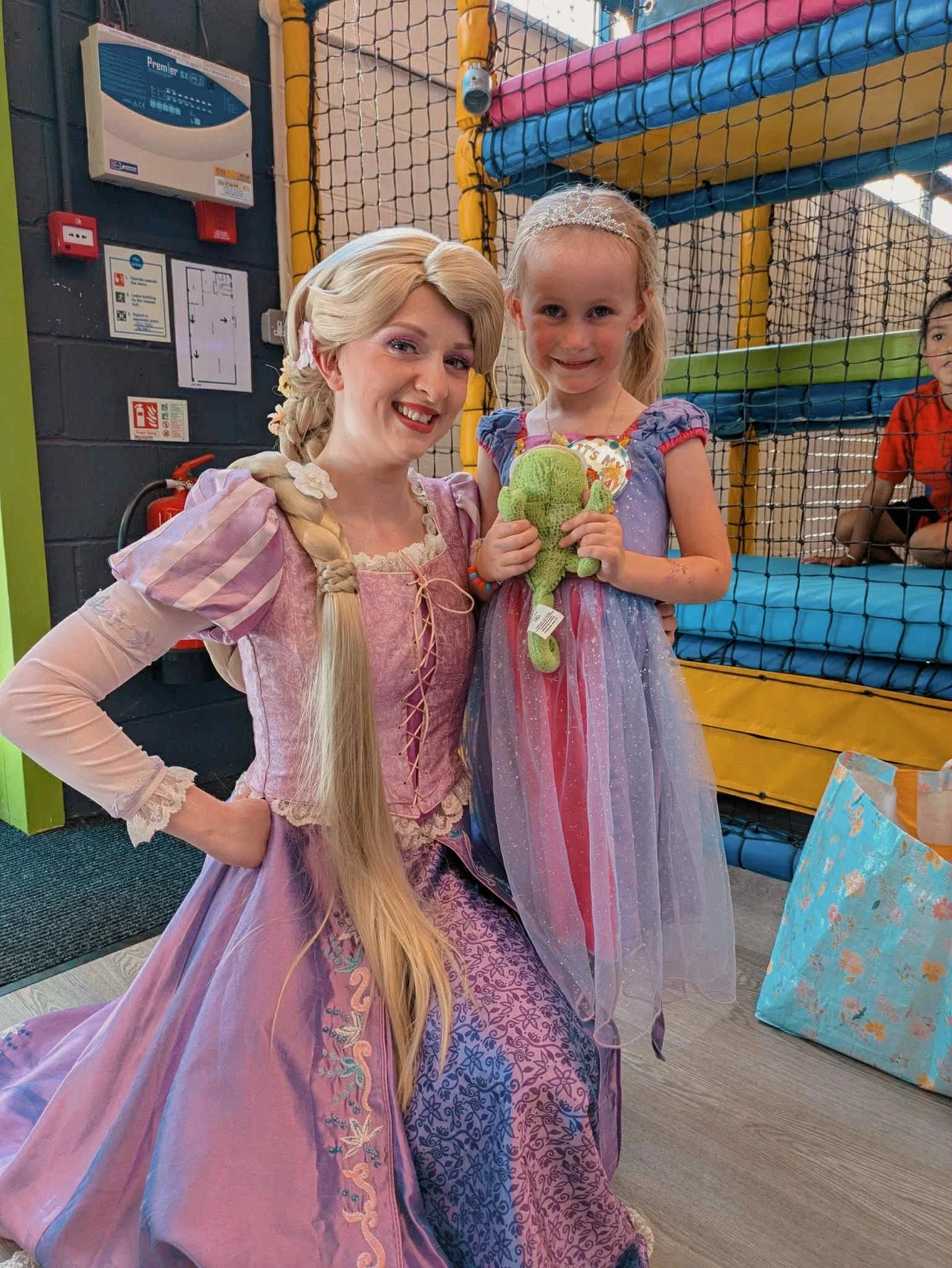 A woman dressed as Rapunzel from Disney's Tangled, with long blonde hair, poses with a young girl in a princess dress at an indoor play area. The girl is smiling and holding a green stuffed toy. The background features colorful play structures and a 