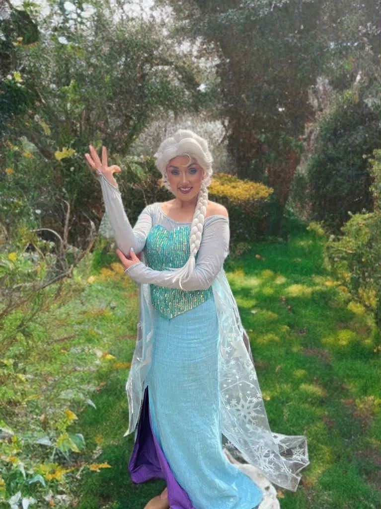 Woman dressed as Elsa from Frozen, standing outdoors in a garden with green bushes and flowers, waving and smiling.