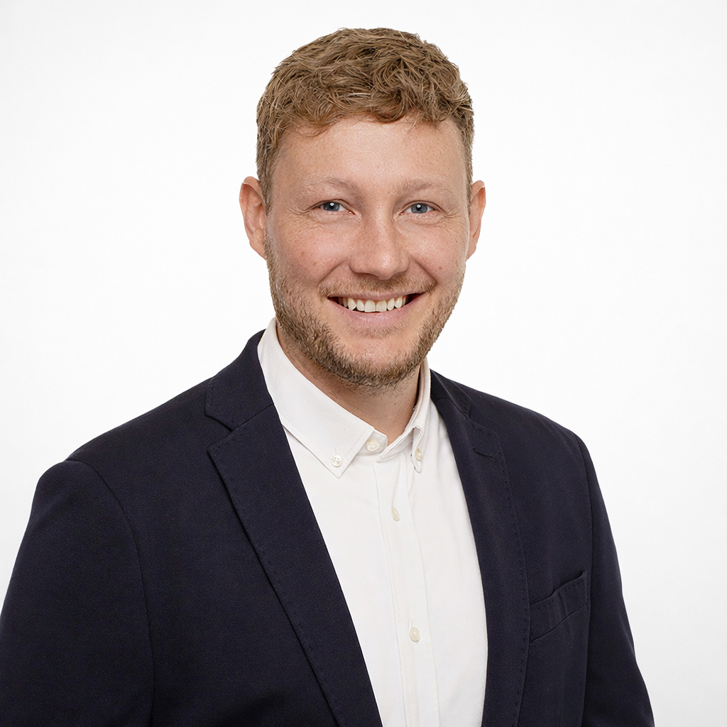 Ein Lächelnder Mann in formeller Kleidung, Weißes Hemd und dunkler Blazer, vor weißem Hintergrund.