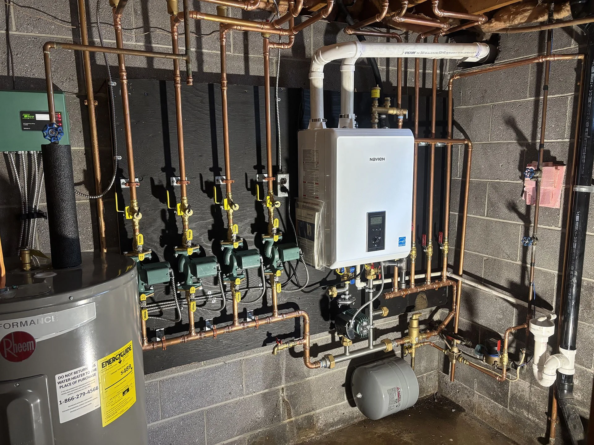 A plumbing setup with interconnected copper pipes, a water heater, and a white Navien tankless water heater installed on a cinder block wall.