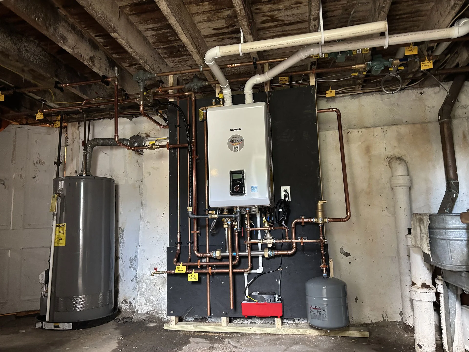 A basement mechanical room featuring a Navien tankless water heater, copper plumbing pipes, a water softener, and a gray expansion tank mounted on a concrete wall.