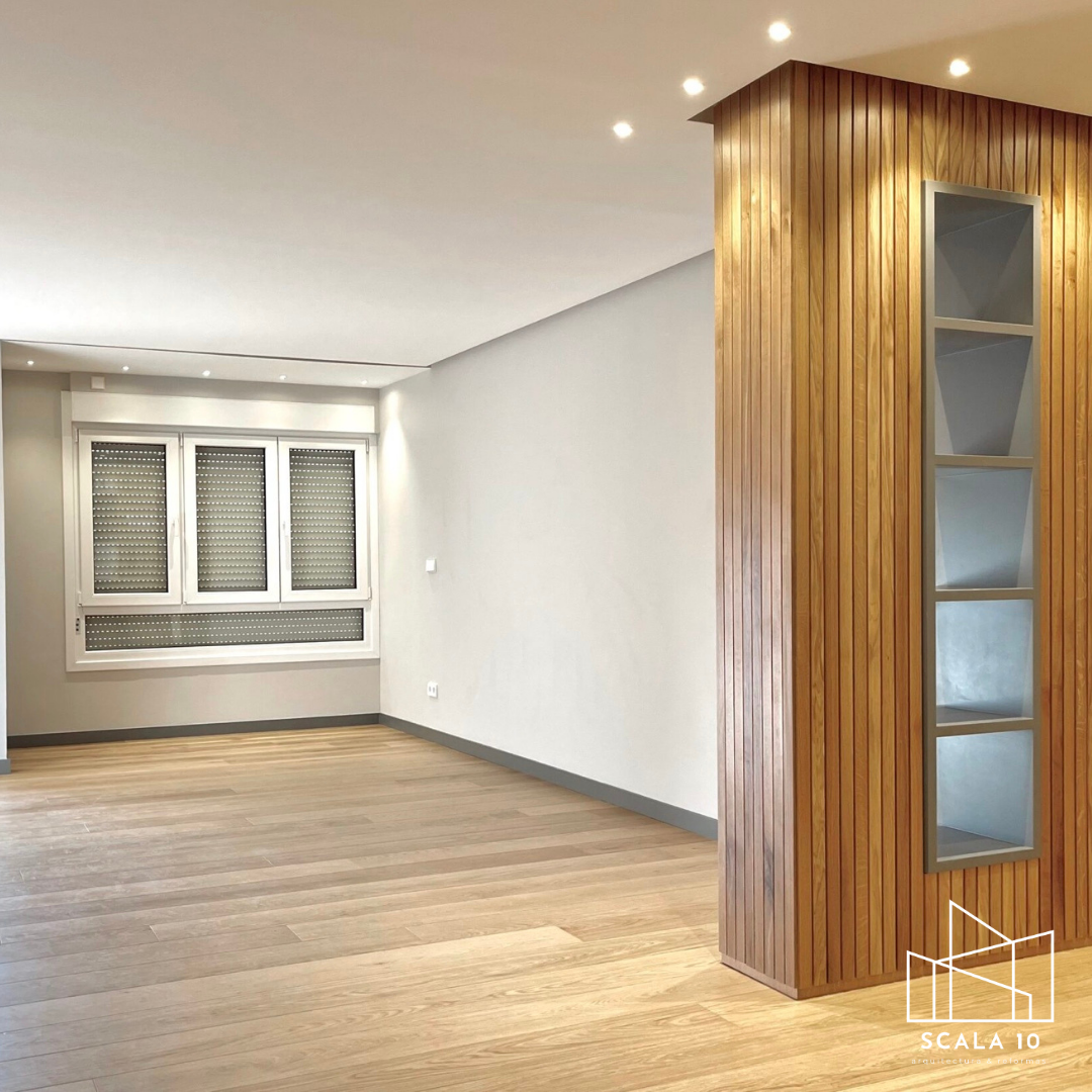 Espacio interior de apartamento o casa con paredes blancas, piso de madera clara, ventana grande con persianas de seguridad, y una estructura de madera con estantes empotrados.