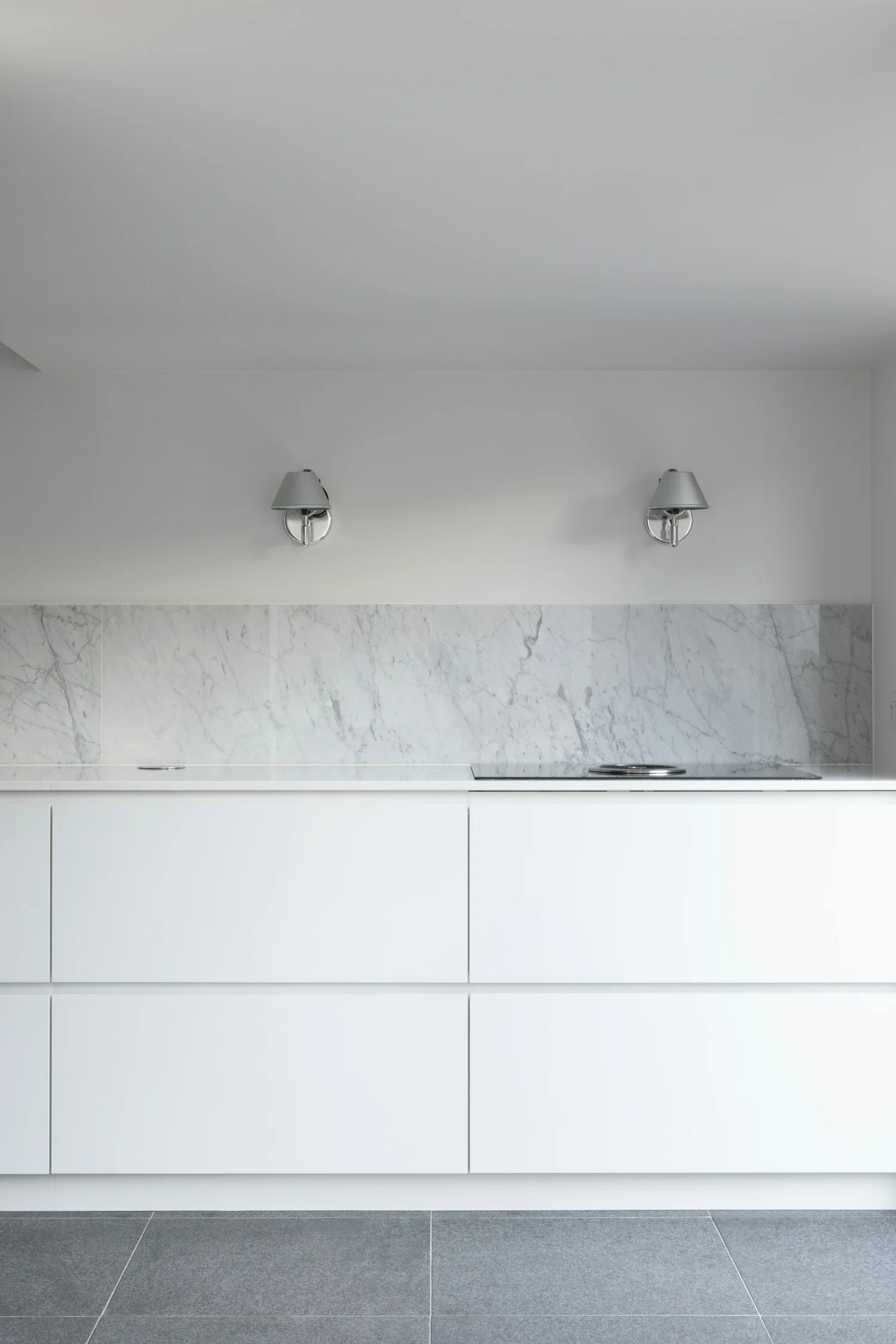 Cocina moderna con gabinetes blancos, encimera de mármol y dos apliques de pared con pantallas plateadas en la pared del fondo.