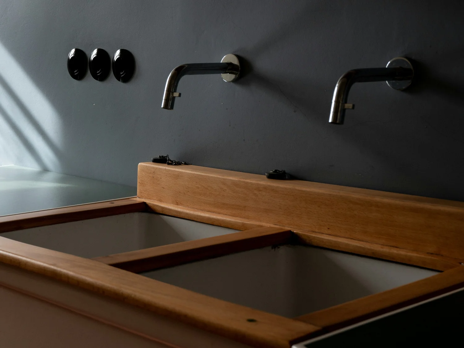 Espejos con luz de espejo en una pared gris claro, con grifos de acero inoxidable y una superficie de madera debajo.