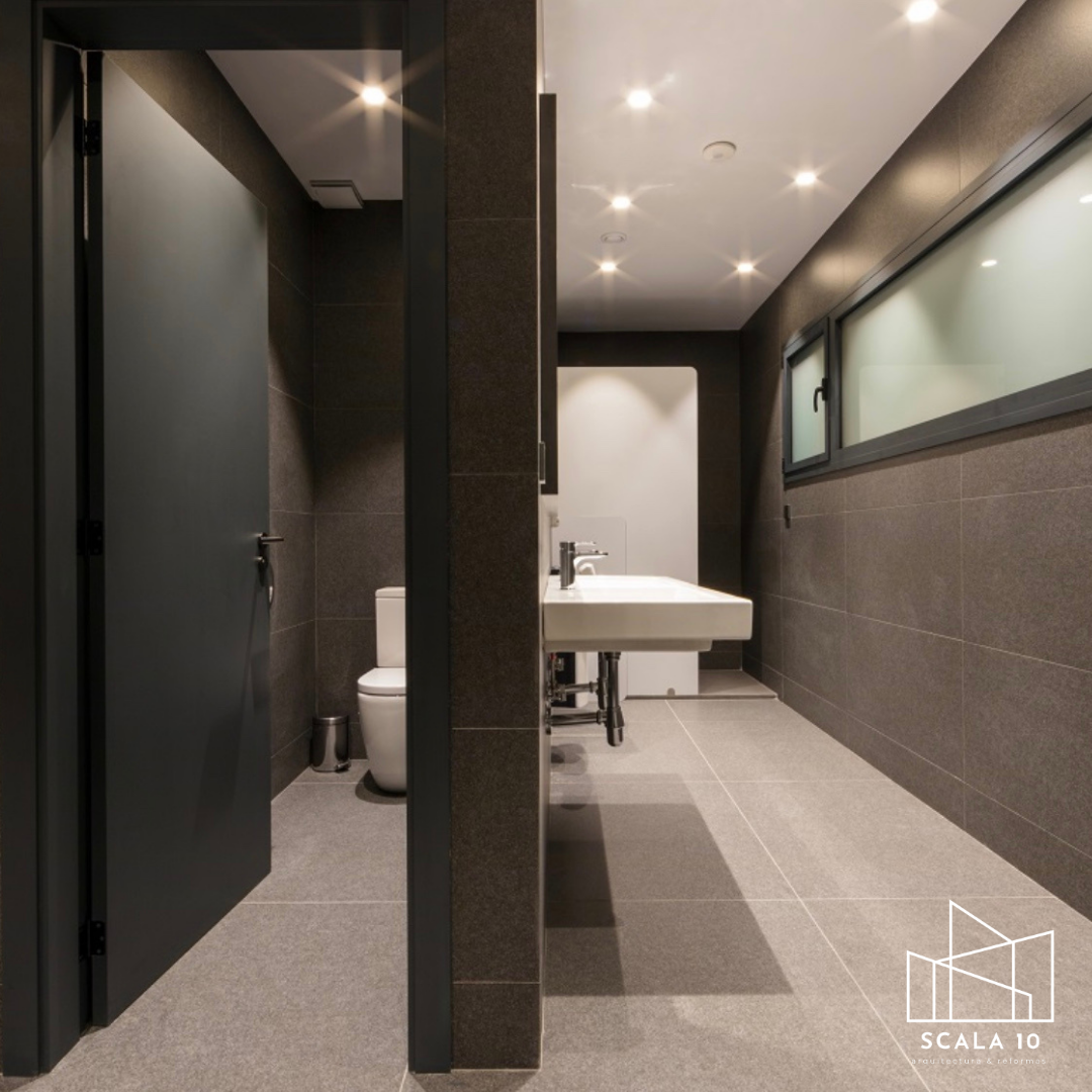 Baño moderno con paredes y piso de baldosas grises, puerta negra, fregadero y ventanas opacas.
