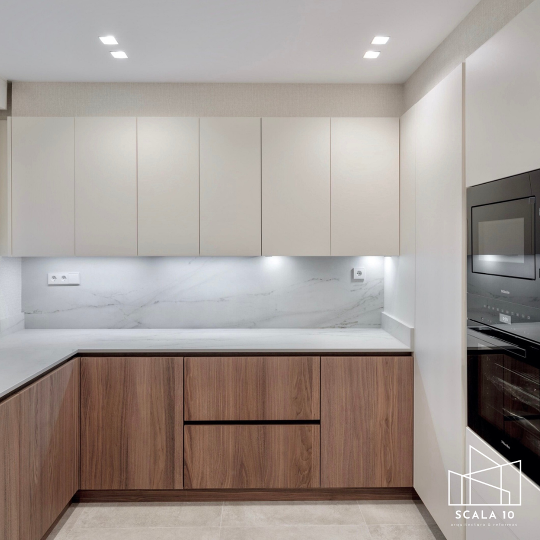 Cocina moderna con gabinetes blancos y de madera, encimera de mármol y electrodomésticos integrados en tonos oscuros.