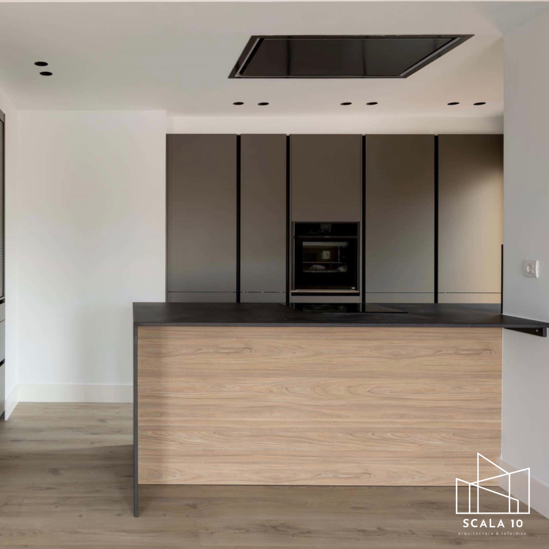 Cocina moderna con isla de madera clara y encimera negra, armarios grises y horno empotrado, suelo de madera y techo con panel central y varias luces pequeñas.