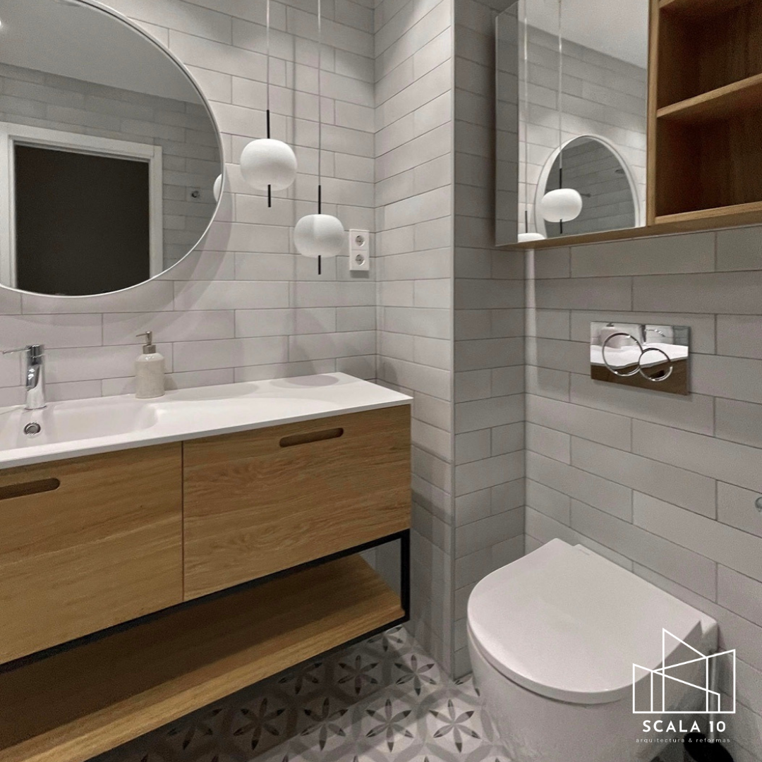 Baño moderno con pared de azulejos blancos y suelo de baldosas con diseño geométrico, espejo redondo sobre un lavabo de madera, iluminación con dos lámparas colgantes blancas, y un inodoro en la esquina derecha, con repisa de madera abierta y enchufes cerca del espejo.