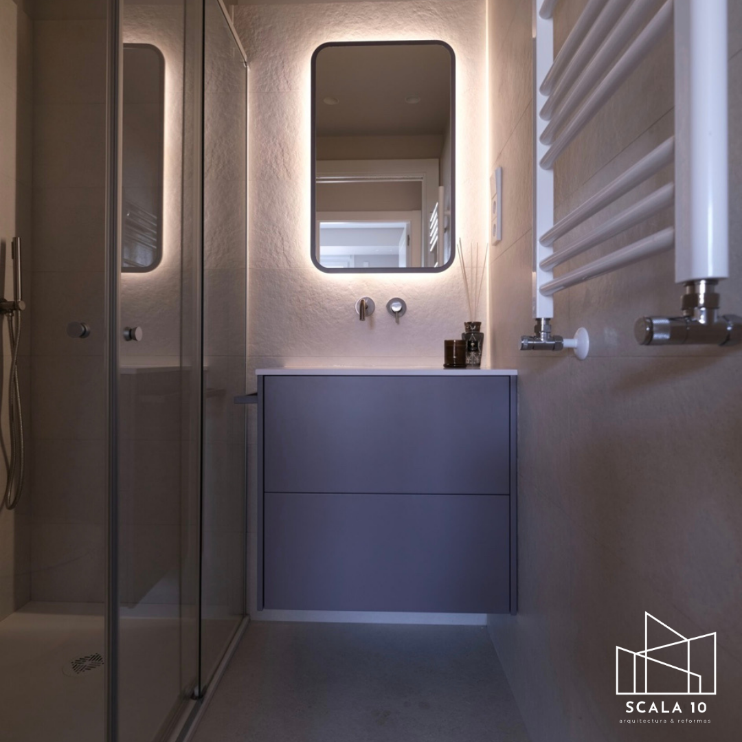 Baño moderno con ducha, espejo rectangular con iluminación de fondo, lavabo gris con lavabo sobre mueble, accesorios de baño y radiador de toallas blanco.