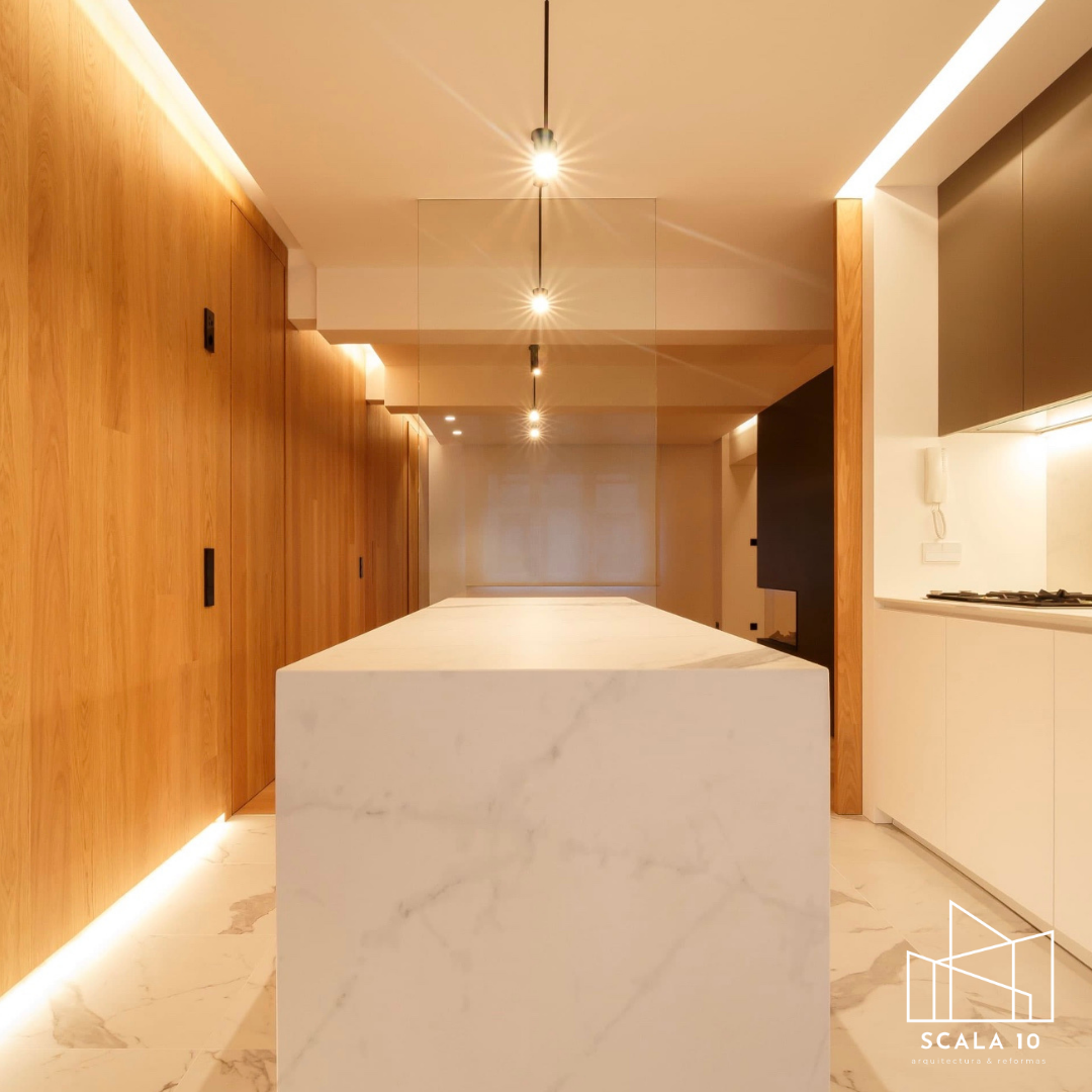 Cocina moderna con isla de mármol blanco, paredes de madera, gabinetes blancos y iluminación empotrada.