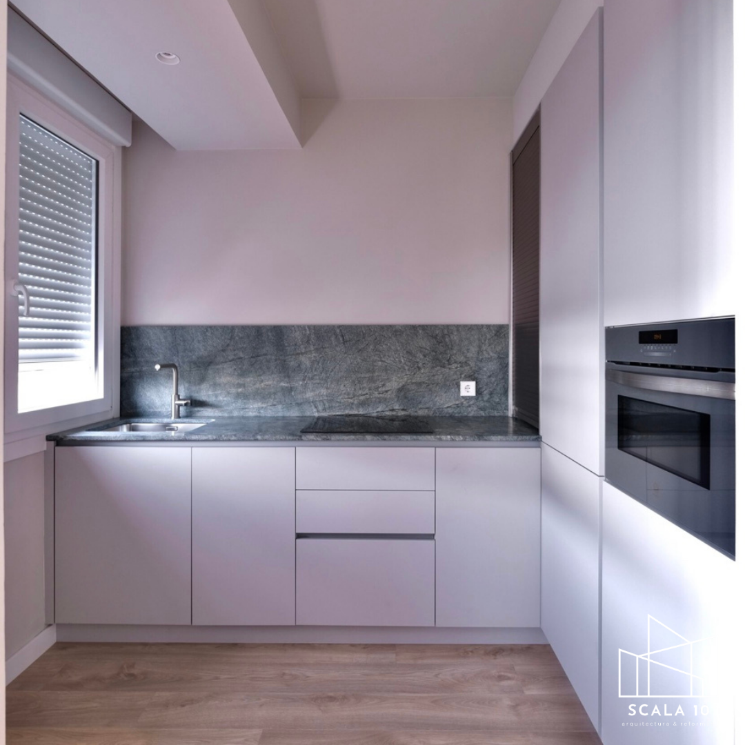 Cocina moderna con gabinetes blancos, encimera de mármol gris oscuro, lavaplatos y horno empotrado, ventana con persiana