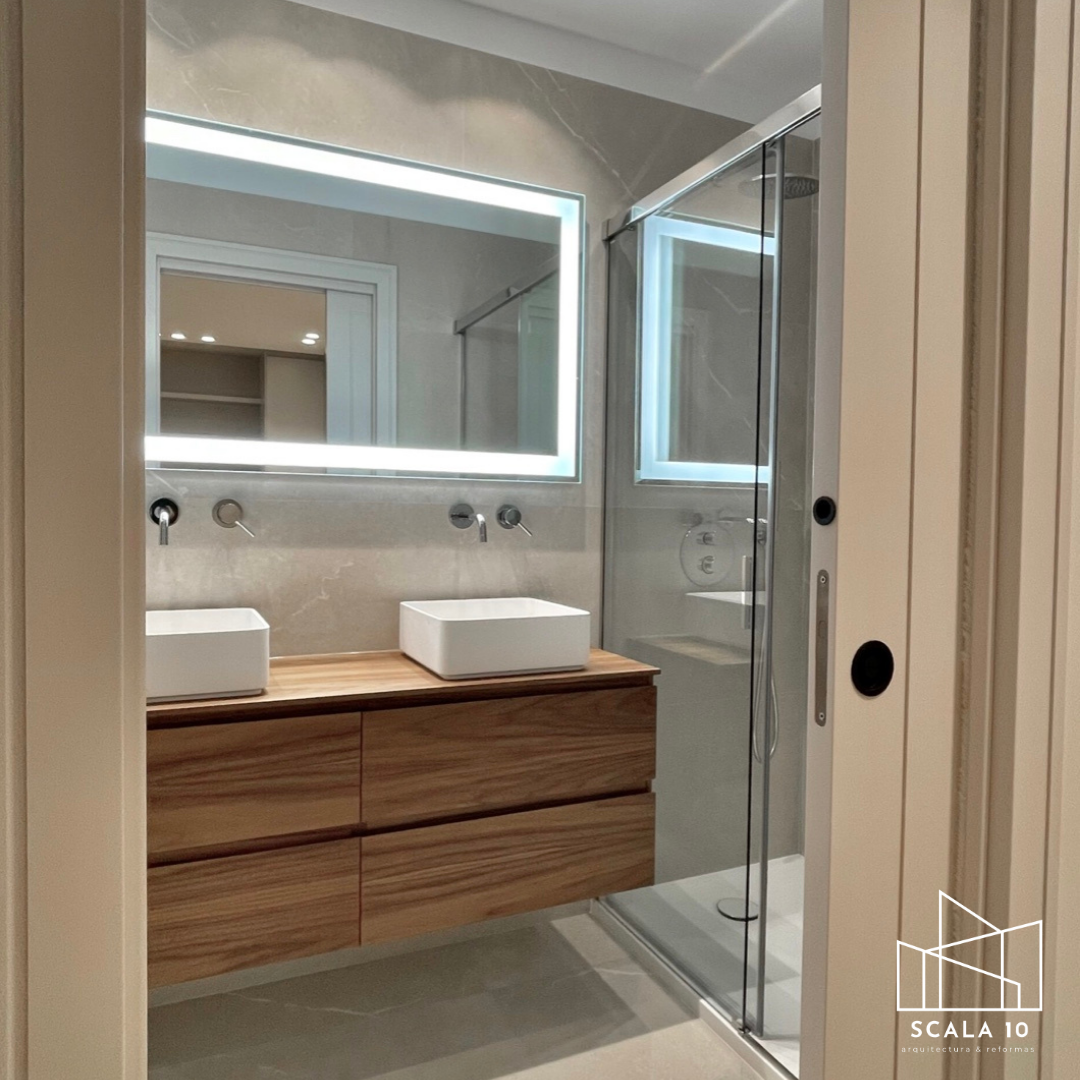 Imagen de un baño moderno con espejo con iluminación LED, lavabo de madera y dos minerales blancos, ducha con puertas de vidrio.