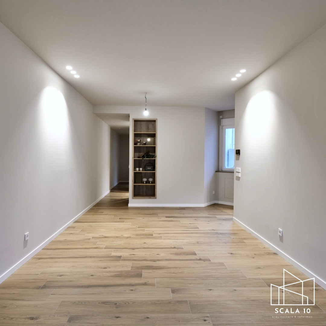 Espacio interior de sala o comedor con paredes blancas y suelo de madera clara, iluminado por luces empotradas en el techo. Ventana en la esquina derecha y estantería abierta al fondo.