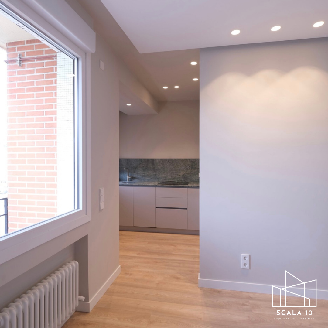 Interior de una habitación con pared blanca, ventana grande con vista a un edificio de ladrillos, piso de madera clara, y en el fondo una pequeña cocina con encimera de granito y gabinetes grises. Se observan focos empotrados en el techo.