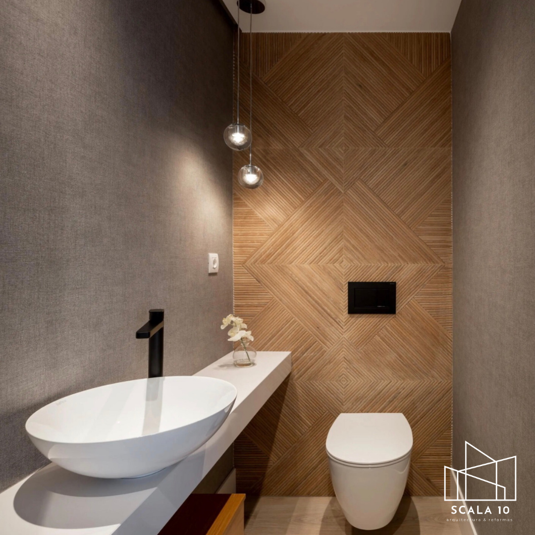 Espacio de baño con pared decorativa de madera, lavabo ovalado en lavabo flotante, lavabo pequeño decorado con flor, inodoro blanco y moderno, con luz suspendida y pared gris de fondo.