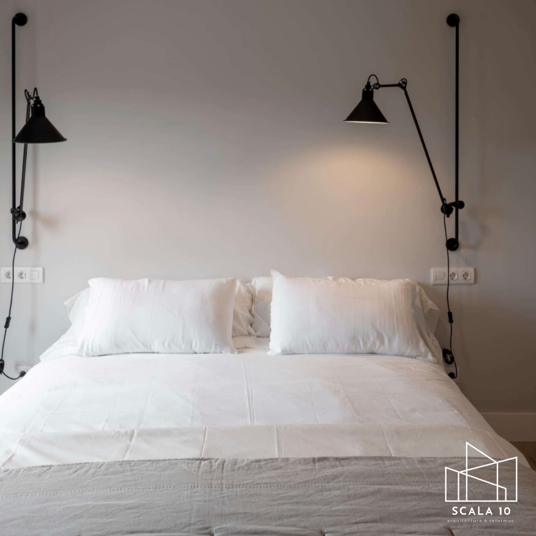 Cama con sábanas blancas y almohadas blancas en un dormitorio moderno con paredes grises. Lámparas de pared negras con brazos articulados a ambos lados de la cama, con tomacorrientes en las paredes.