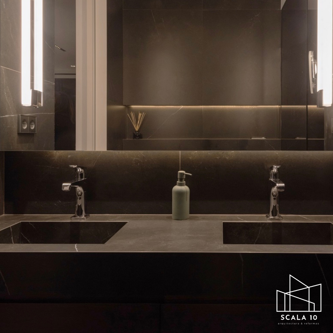 Lavabo doble de piedra negra con dos grifos y un dispensador de jabón en medio, espejo grande con iluminación, y paredes de piedra negra en un baño moderno.