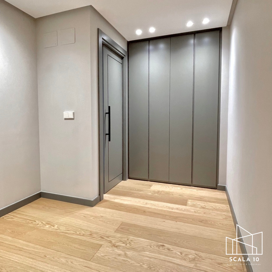 Vista de una habitación moderna con paredes grises, suelo de madera clara, un armario empotrado gris y una puerta gris con manija negra, iluminación en el techo y una toma de corriente en la pared.